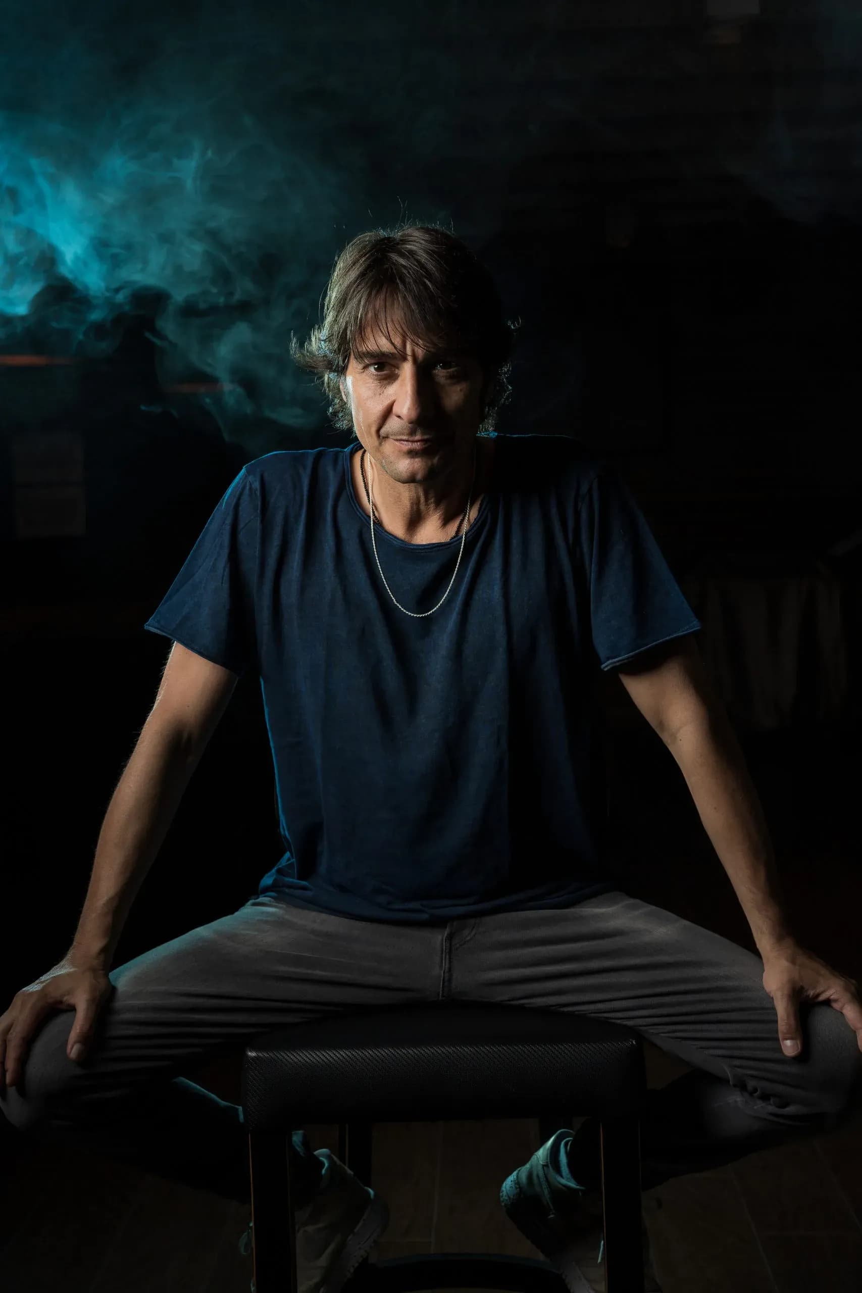 Abel Almena posando sentado de cuerpo entero con camiseta azul, retrato promocional sobre fondo oscuro con neblina escénica