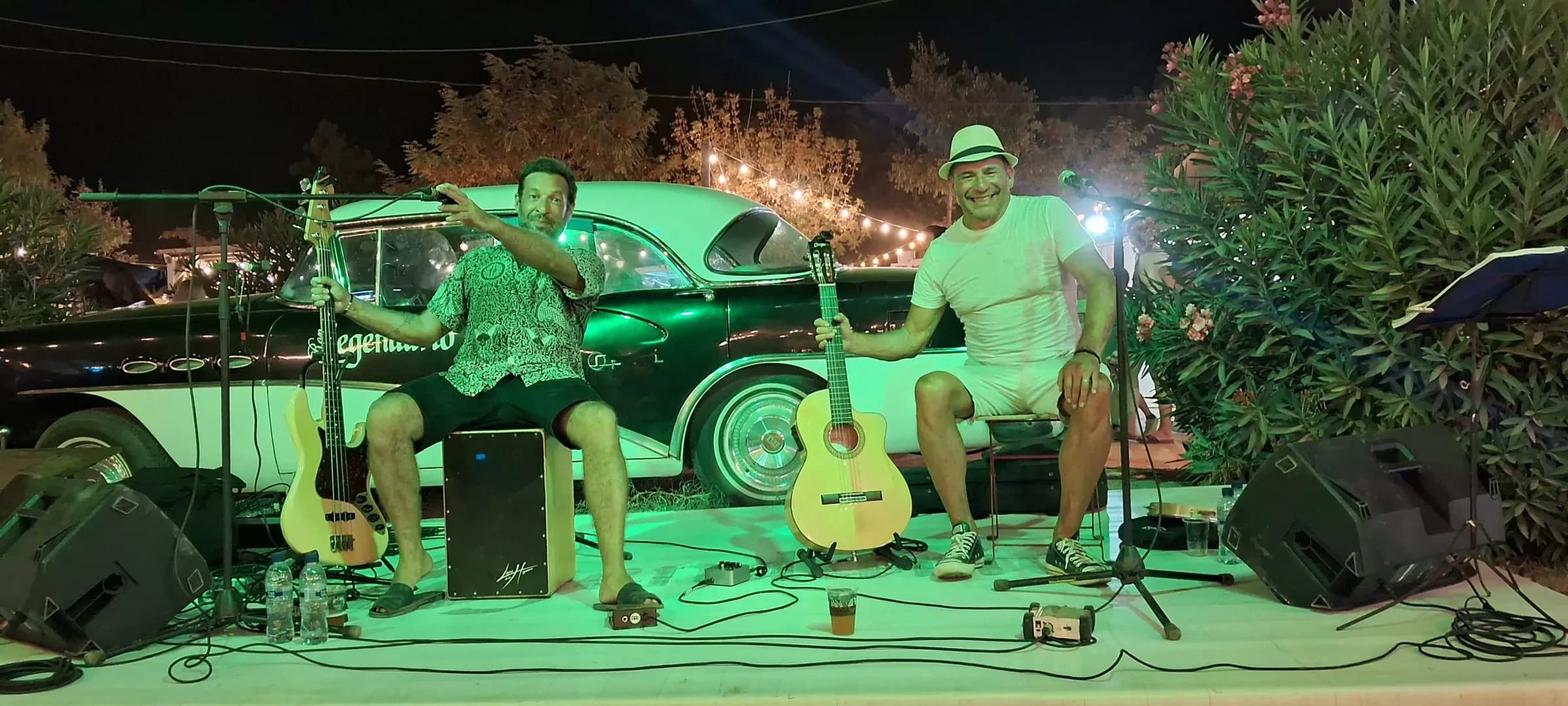 Dúo Barreto y Cía actuando sobre escenario con guitarras frente a un coche americano clásico, ambiente vintage con luz verde