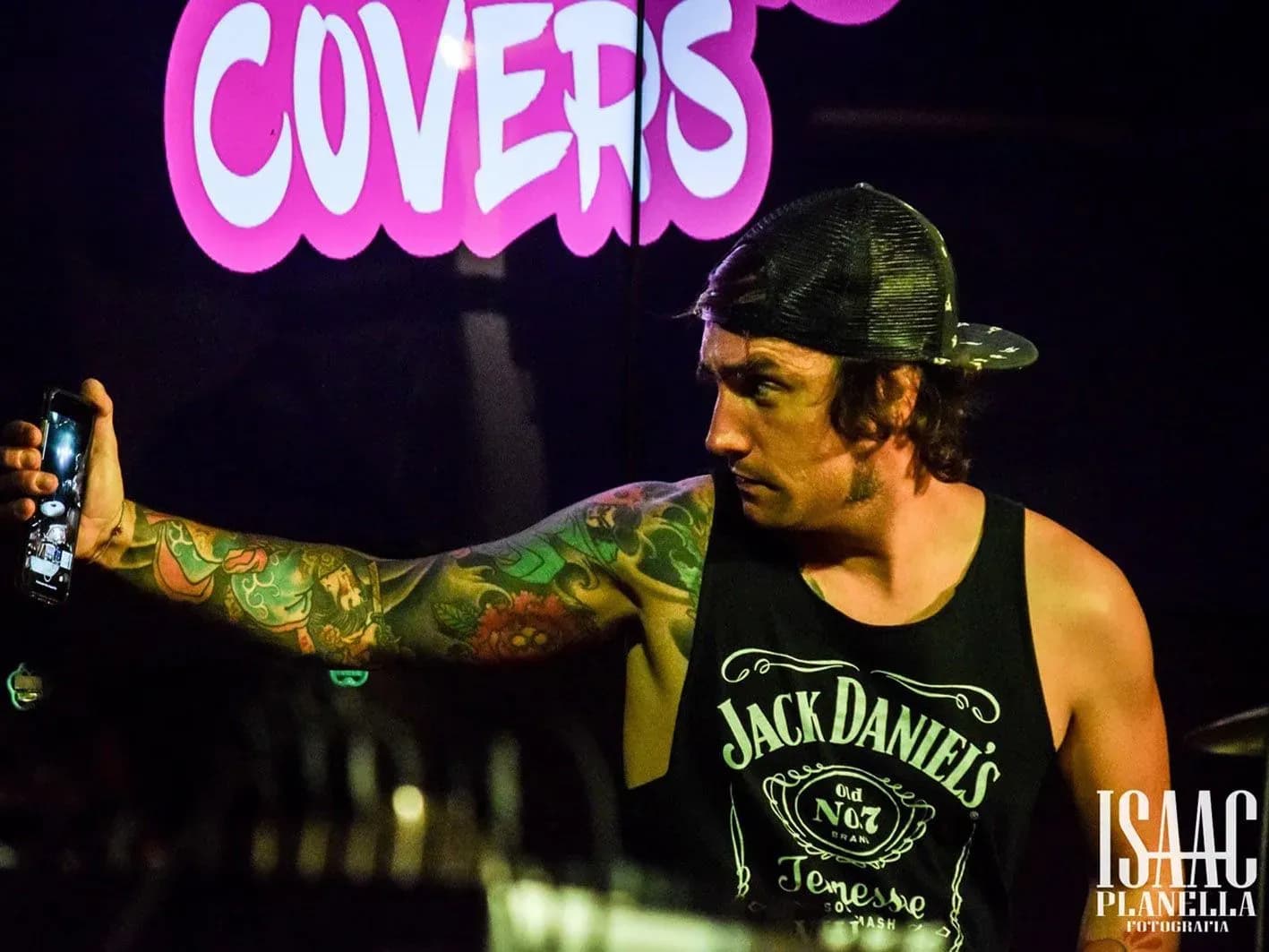 Cantante tatuado de Big Mouthers con camiseta Jack Daniels grabando con móvil ante neón rosa Covers en sala de conciertos