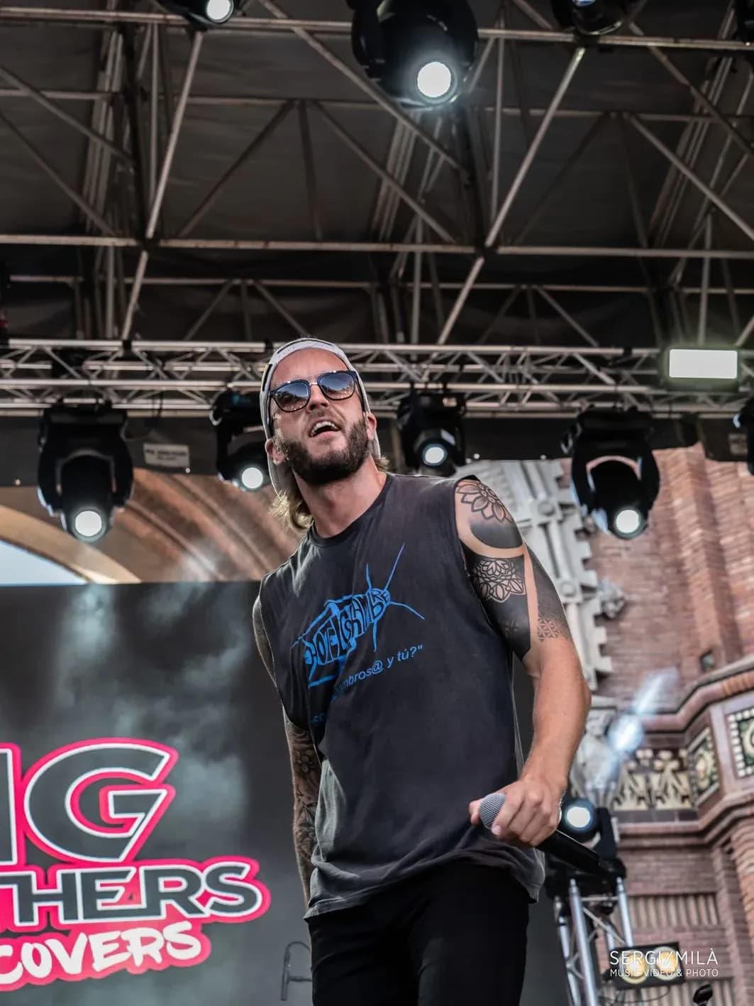 Cantante de Big Mouthers con gorra y gafas de sol sobre escenario al aire libre con tatuajes en el brazo