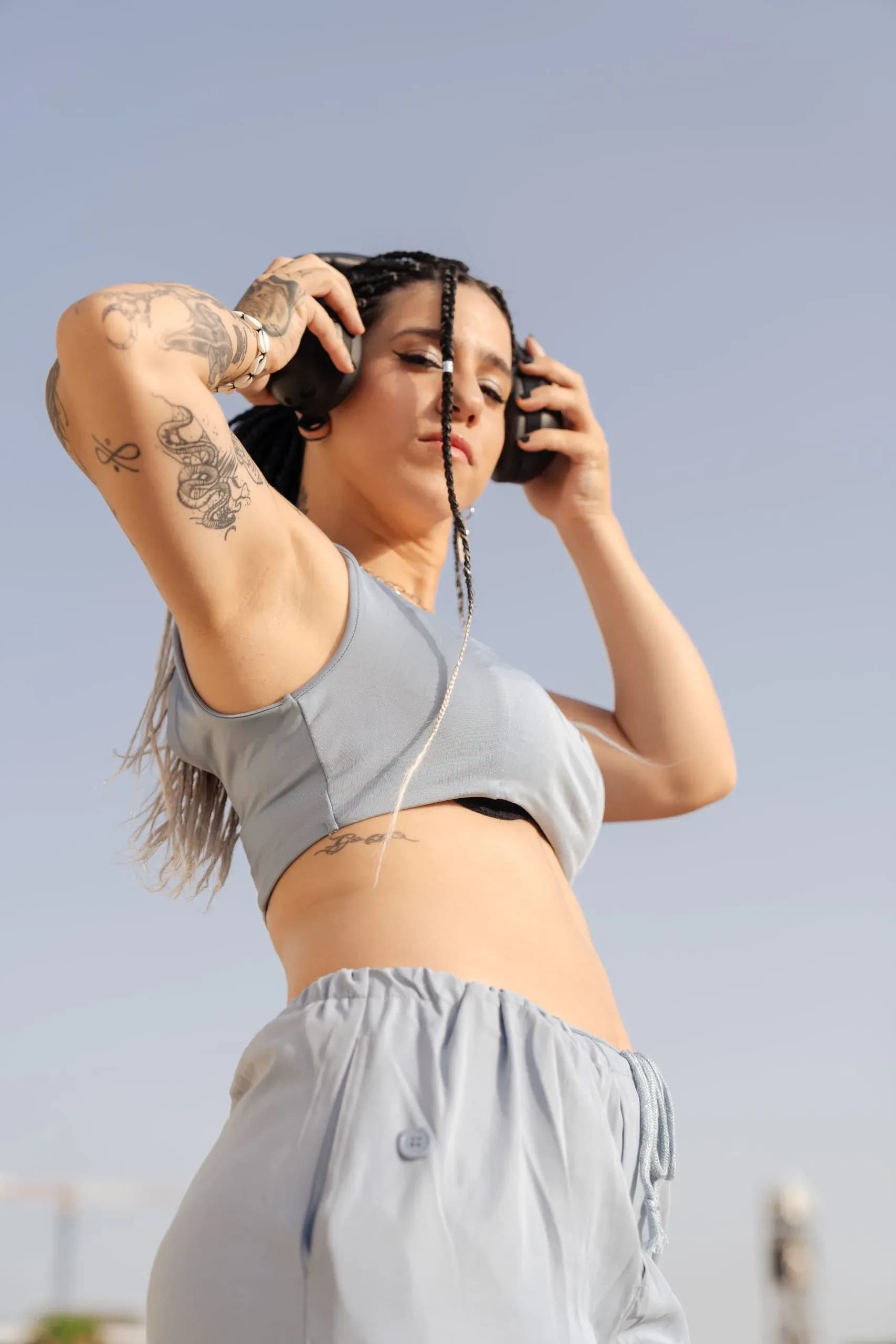 Carla Nisha con top gris y brazos tatuados sosteniendo los auriculares bajo cielo abierto, retrato urbano de la DJ catalana
