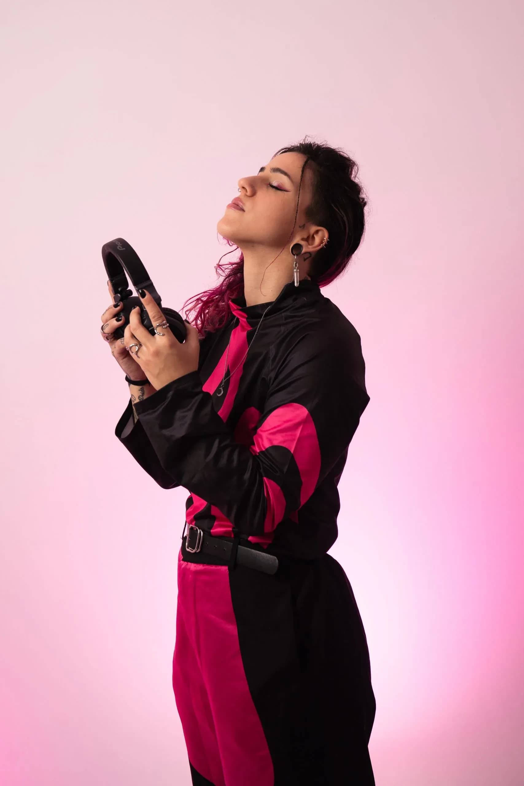 Carla Nisha con chaqueta negra y fucsia sosteniendo auriculares sobre fondo rosa de estudio, sesión promocional de DJ urbana