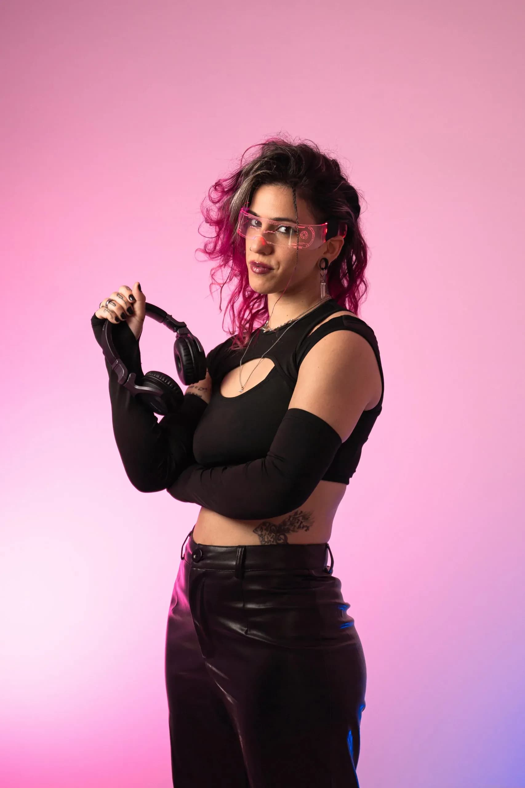 Carla Nisha con gafas LED rosa neón y pantalón de cuero sujetando auriculares, imagen futurista de DJ de afrobeat y reggaeton