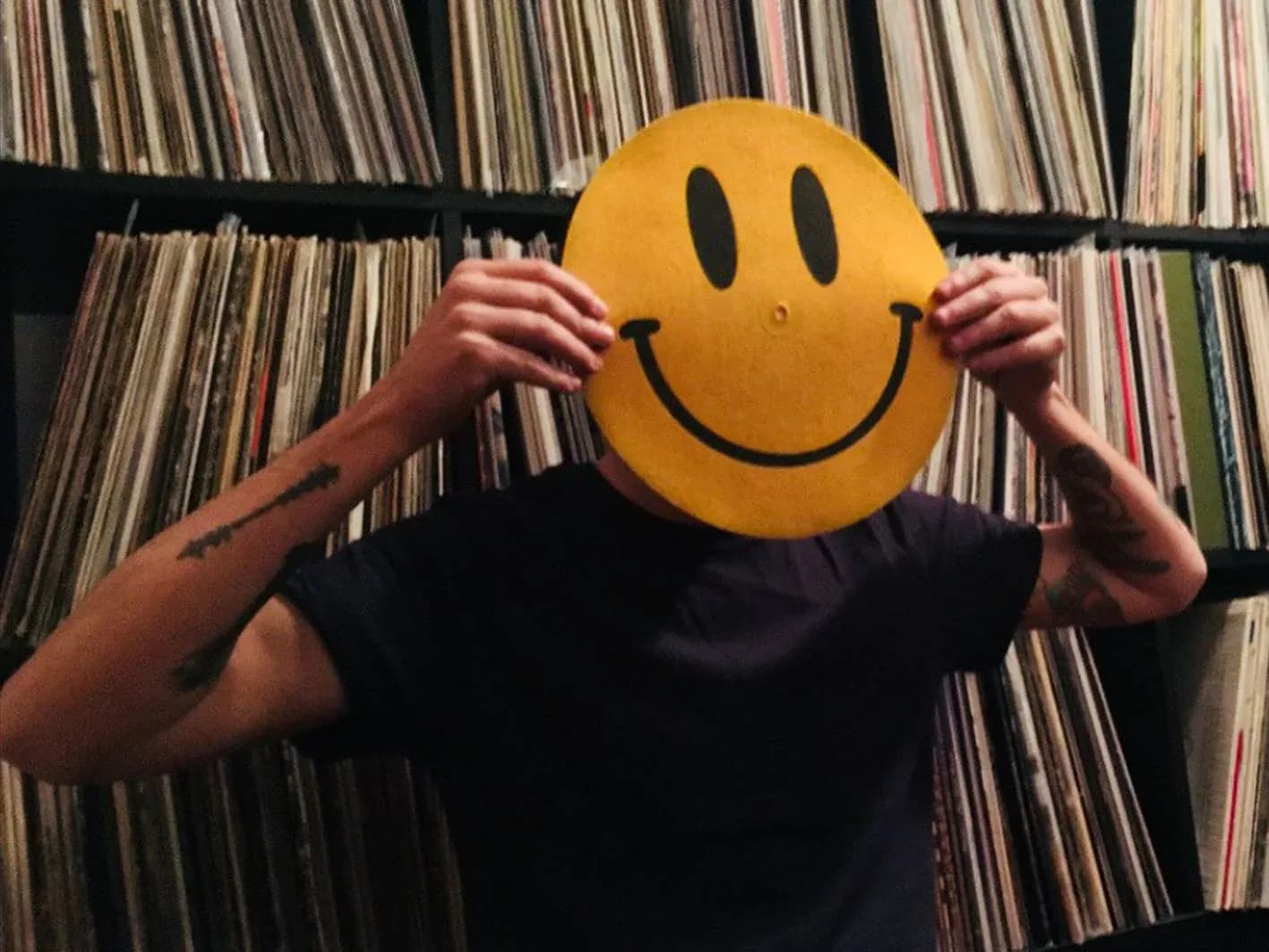 Persona con brazos tatuados sosteniendo un vinilo con cara sonriente amarilla ante estanteria llena de discos