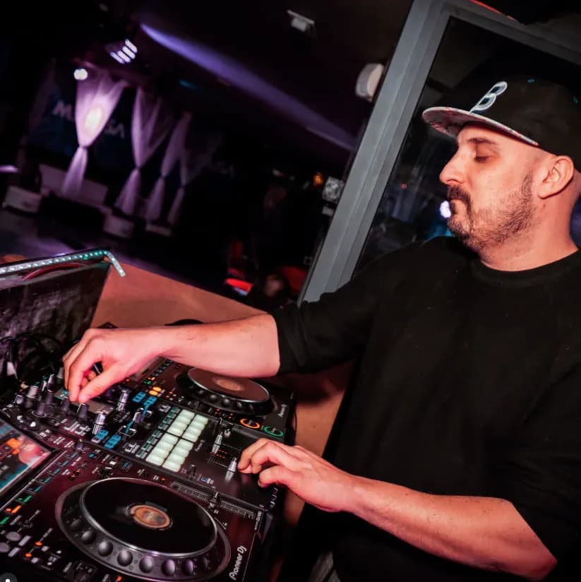 DJ Cruz de Luca con gorra Brooklyn y sudadera negra manipulando mesa Pioneer DJ en cabina iluminada de morado