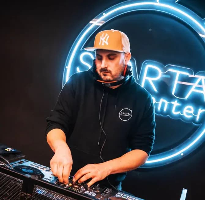 DJ Cruz de Luca con gorra NY naranja y sudadera Sparta Center mezclando ante neón circular azul Sparta en sala