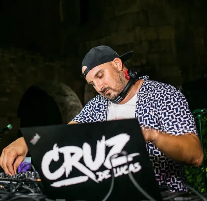 DJ Cruz de Luca con gorra y camisa de cuadritos tras cartel Cruz de Luca, sesión nocturna junto a muro de piedra