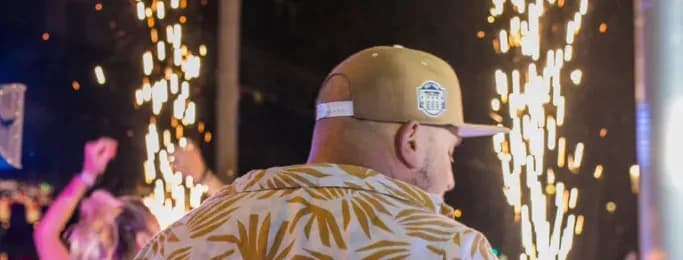 DJ Cruz de Lucas de espaldas con gorra y camisa estampada frente a chispas pirotecnicas en evento nocturno