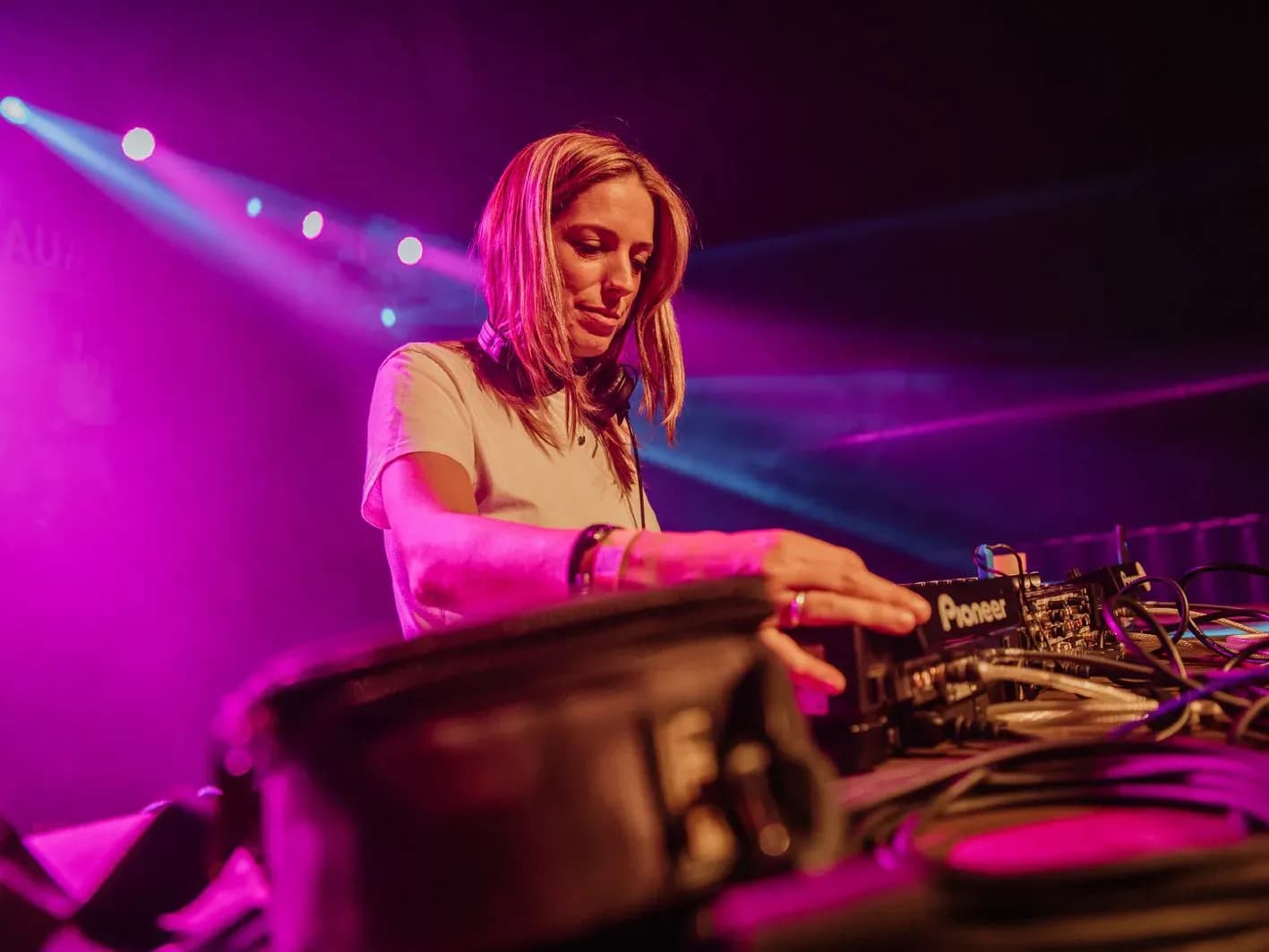 DJ Enka con melena rubia pinchando bajo focos magenta y rayos de iluminación azul en escenario de festival