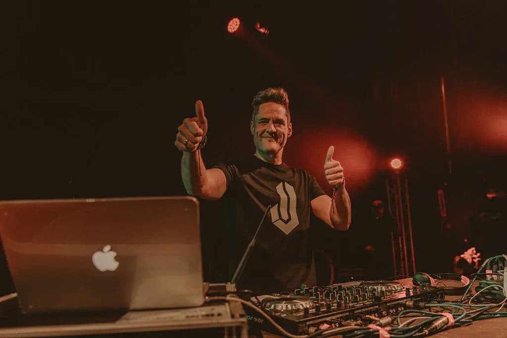 DJ Iván Jiménez levantando ambos pulgares al público desde la cabina con MacBook y mesa Pioneer en concierto