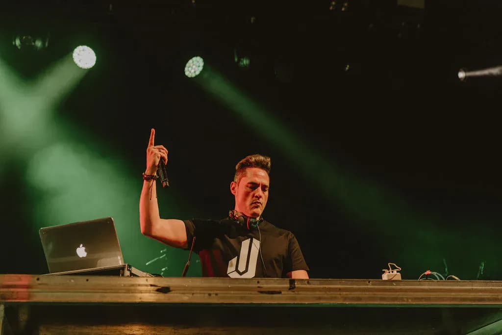 DJ Ivan Jimenez pinchando en escenario con luces verdes, camiseta negra con logo J y portatil Apple