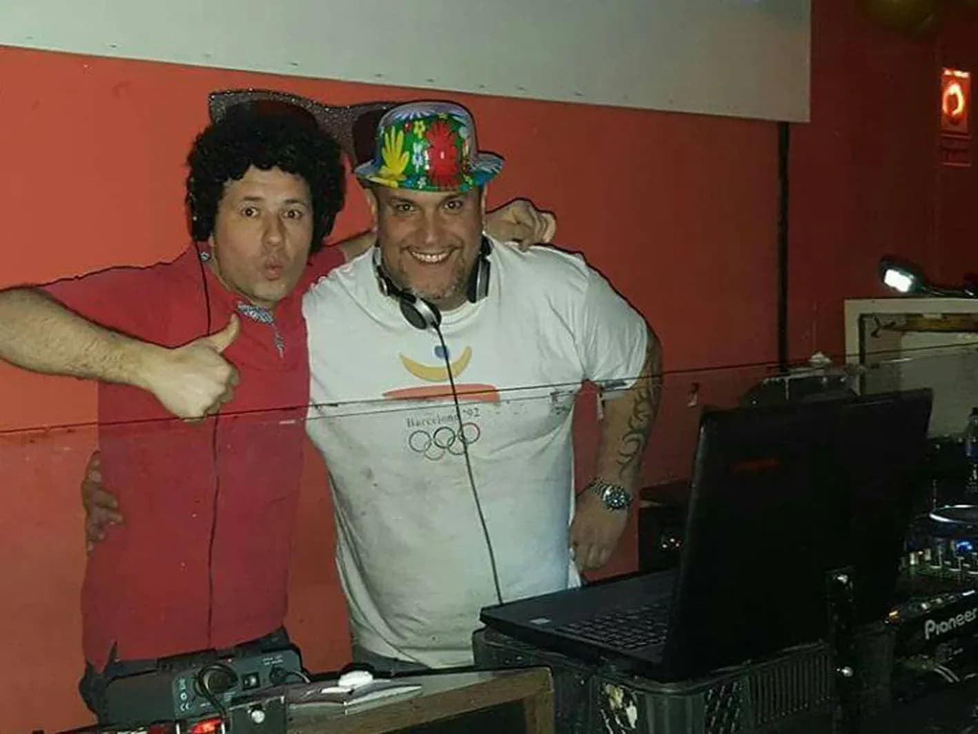 DJ Jordi con sombrero estampado y camiseta Barcelona 92 junto a amigo con peluca afro y gafas gigantes en cabina
