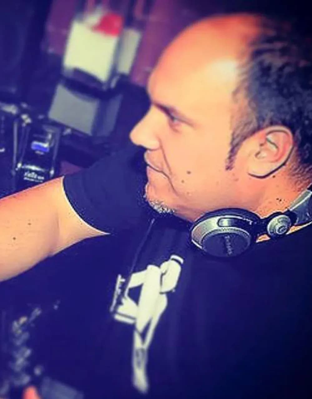 DJ Jordi de perfil con auriculares Pioneer y camiseta negra manejando la mesa en cabina con luz cálida tenue