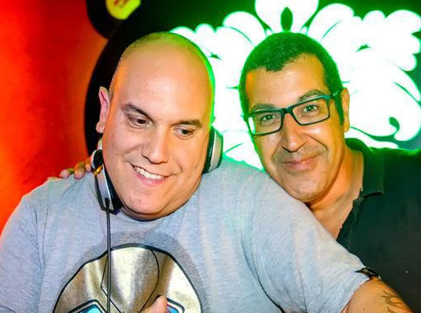 DJ Jordi sonriente con auriculares junto a colega con gafas frente a luminoso floral verde y rojo en club nocturno