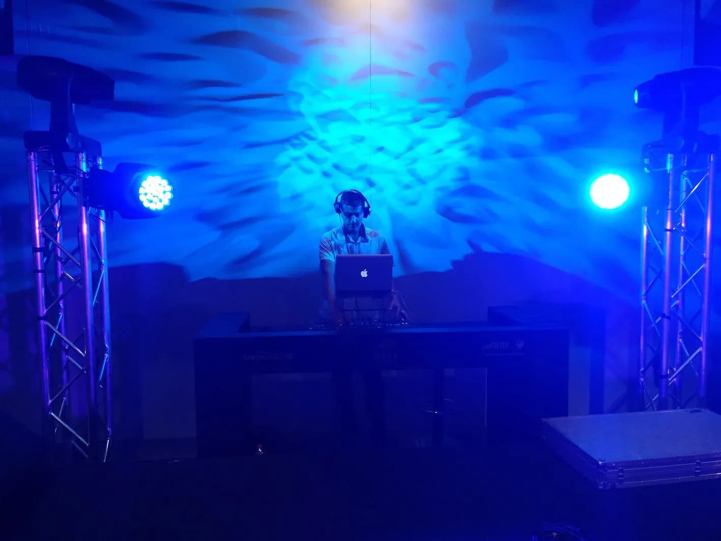 DJ Pablo con auriculares mezclando en portatil sobre cabina iluminada por focos azules y verdes en evento corporativo