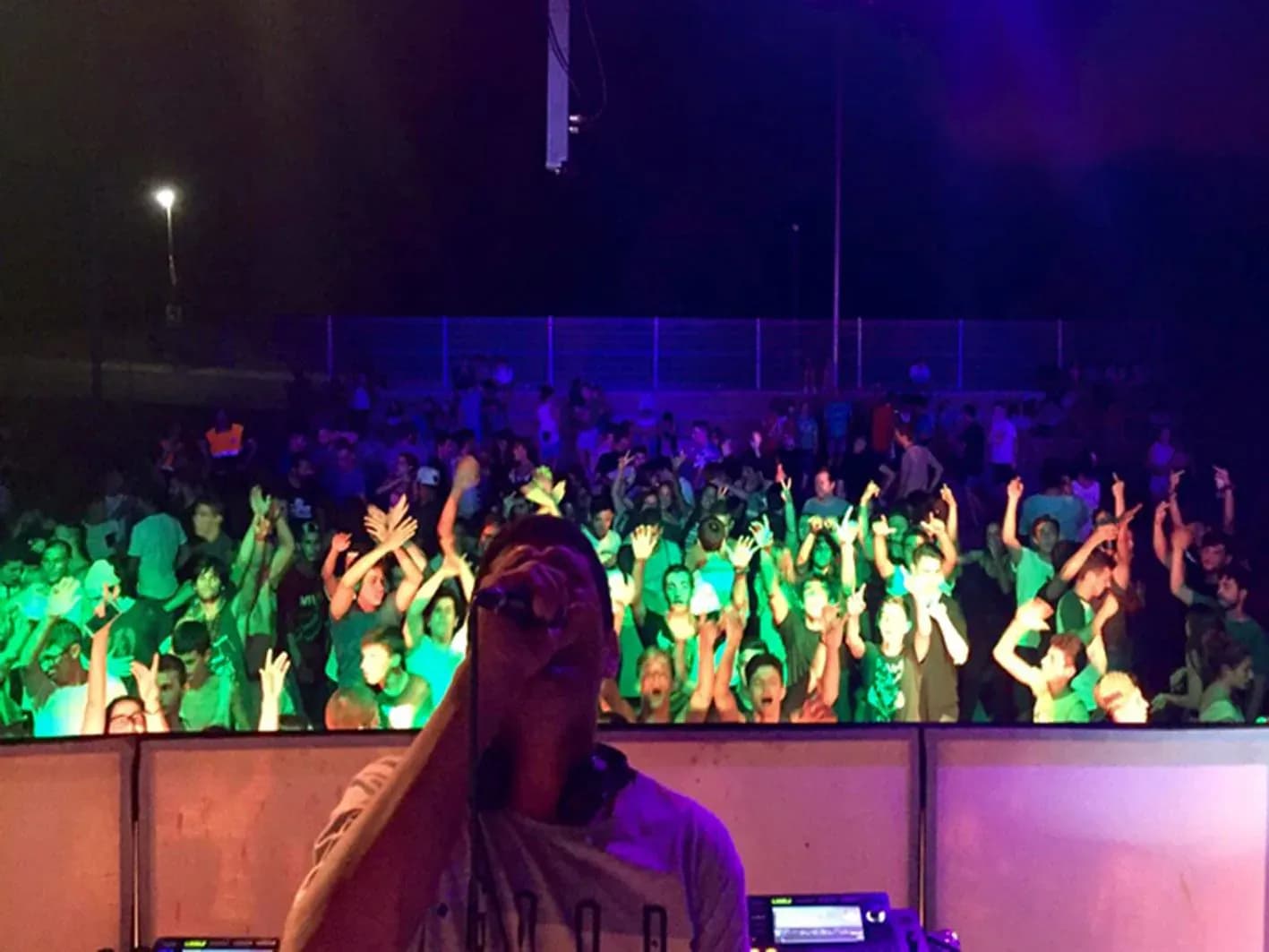 DJ Pablo de espaldas con microfono ante multitud iluminada con luz verde y manos en alto en concierto al aire libre