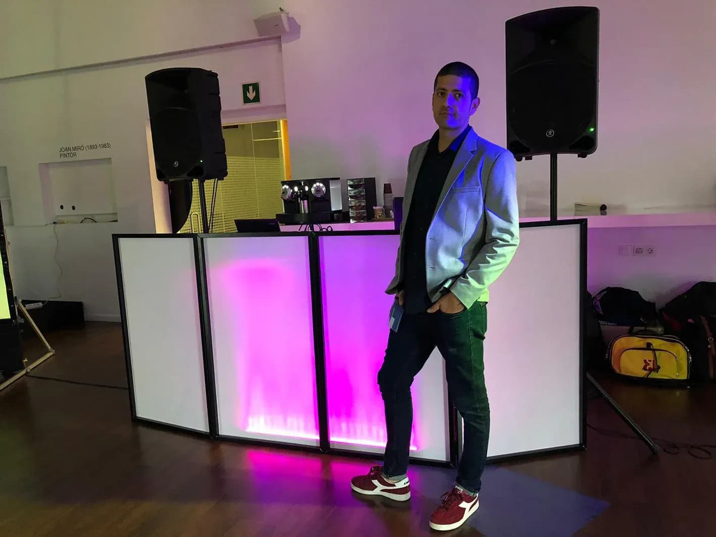 DJ Pablo con americana gris posando frente a cabina iluminada en rosa con dos altavoces y pantallas blancas