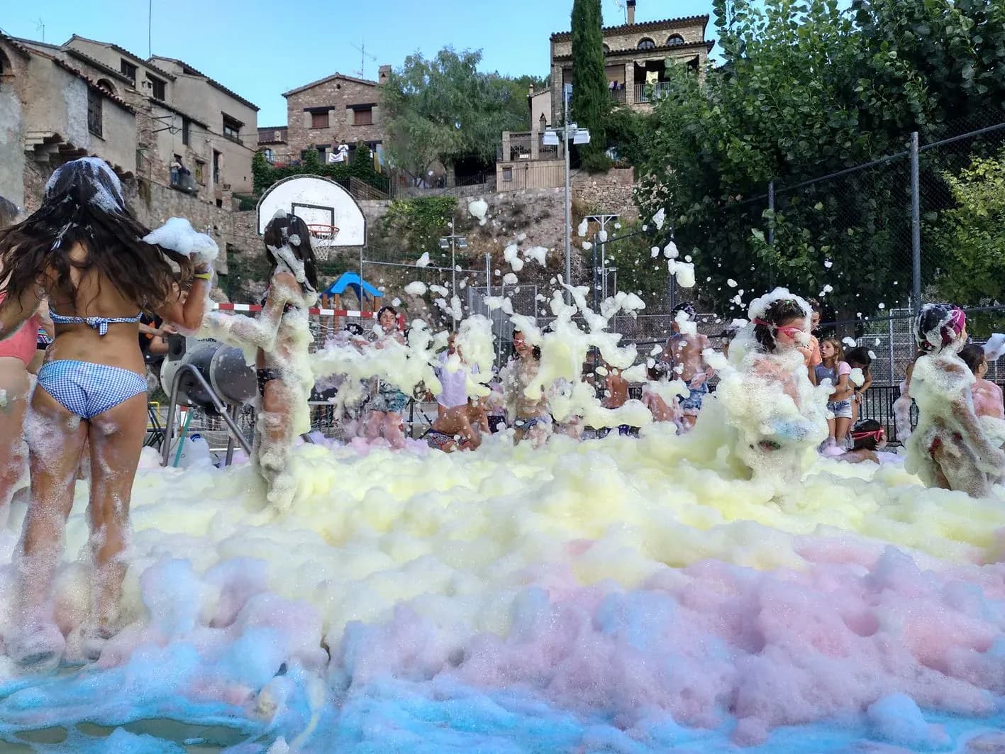 Pista deportiva inundada de espuma de colores con bañistas jugando junto a casas de piedra del pueblo al fondo