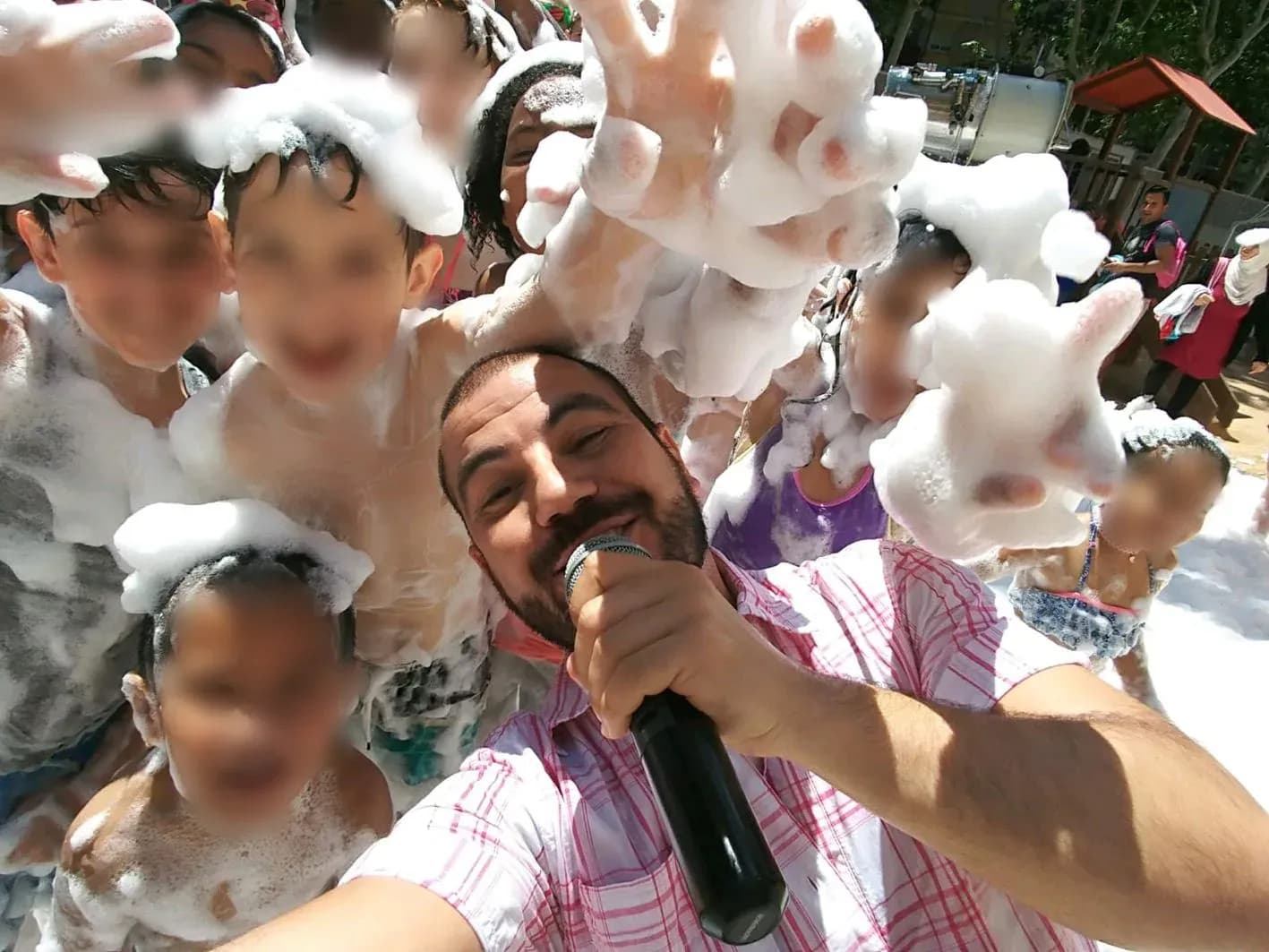 Animador con micrófono cantando rodeado de niños llenos de espuma en fiesta infantil al aire libre durante el verano