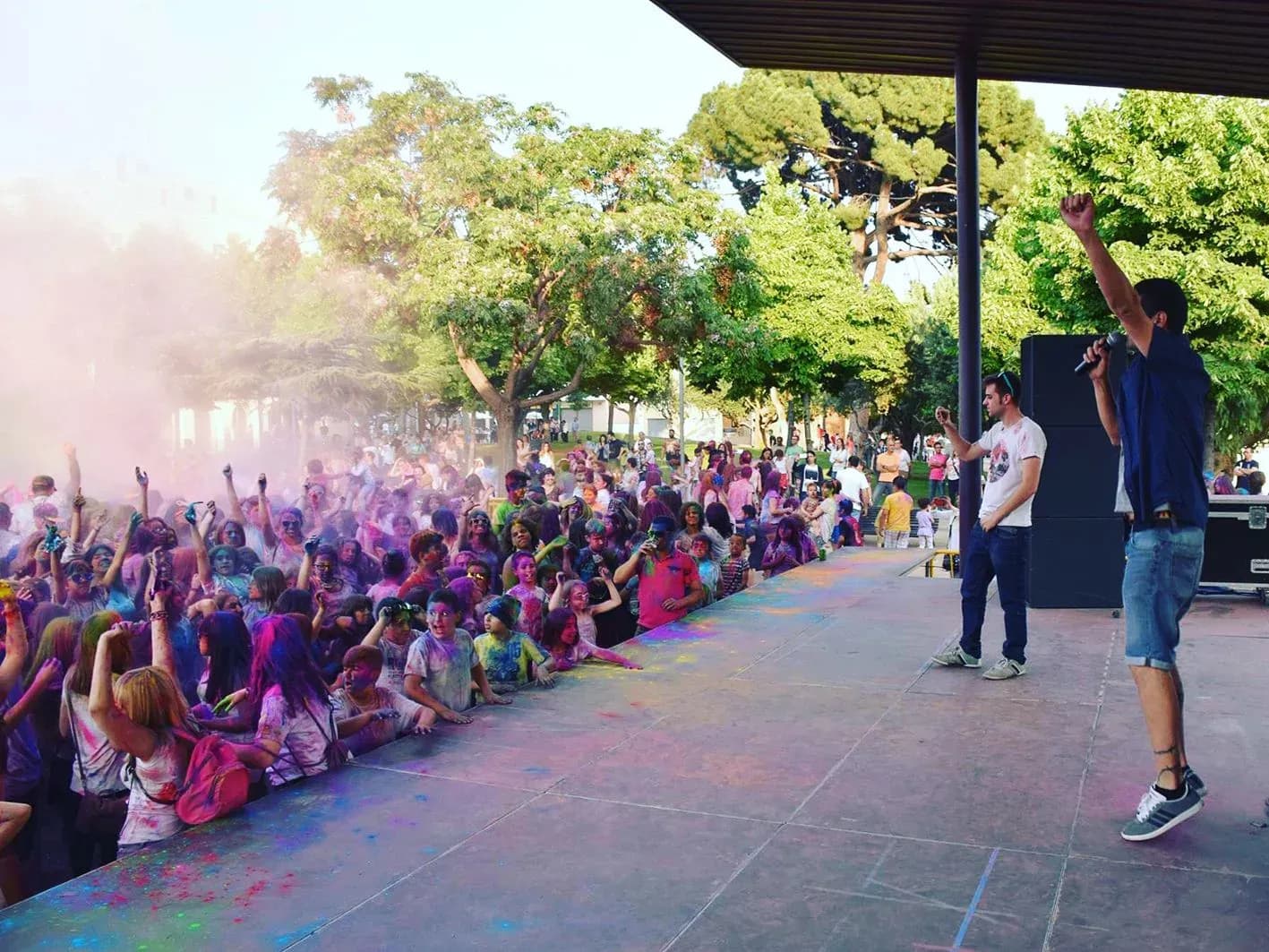 Speakers animando al público con polvos Holi de colores sobre escenario al aire libre con árboles y altavoces al fondo