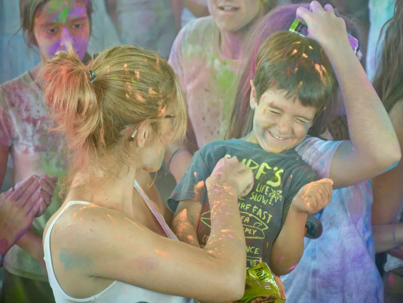 Animadora teñida de Holi entregando bolsita de polvos a un niño sonriente entre nube de colores en fiesta de pueblo