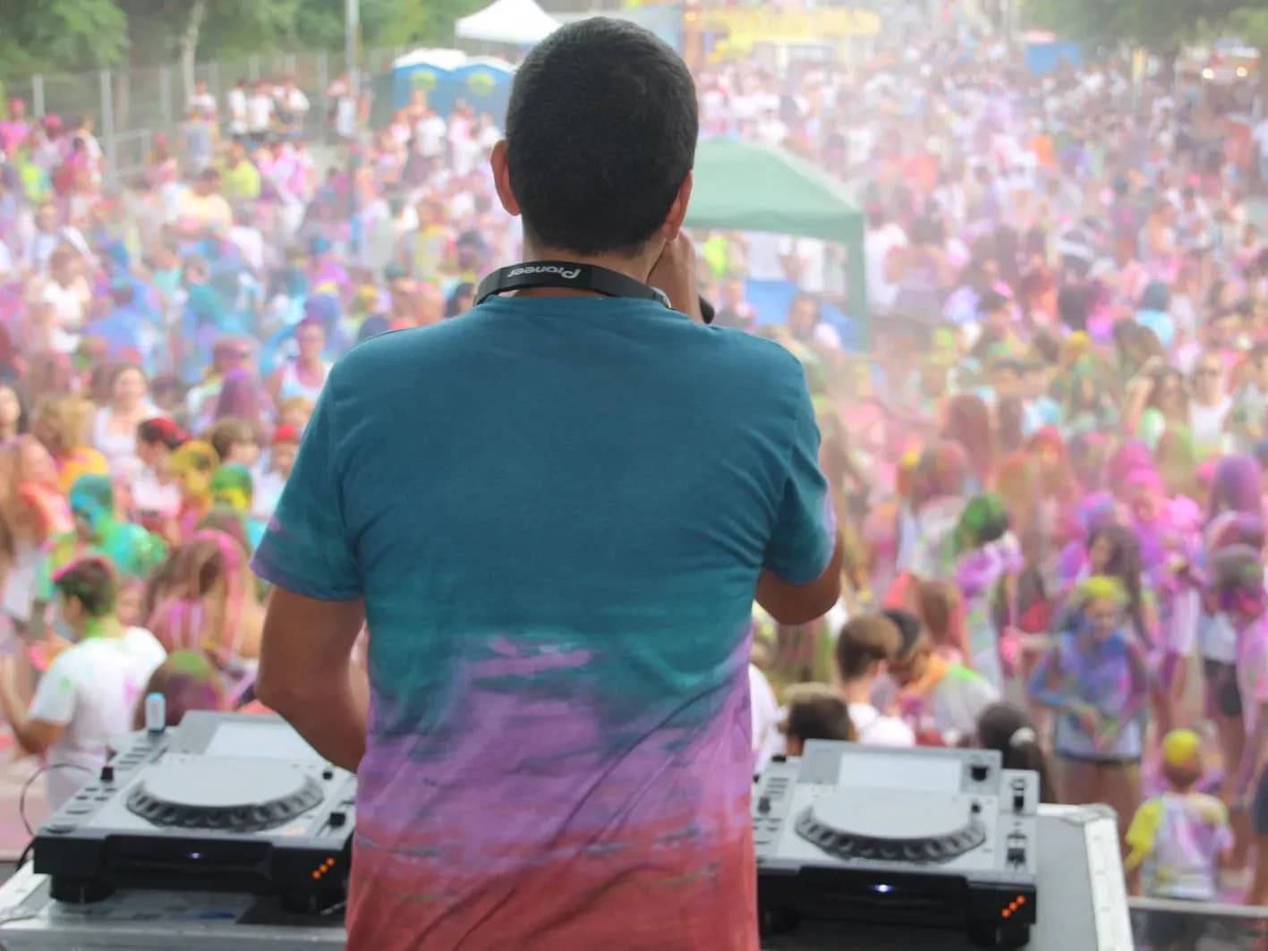 DJ de espaldas con camiseta teñida de polvos Holi mezclando con CDJs Pioneer frente a multitud llena de color en fiesta