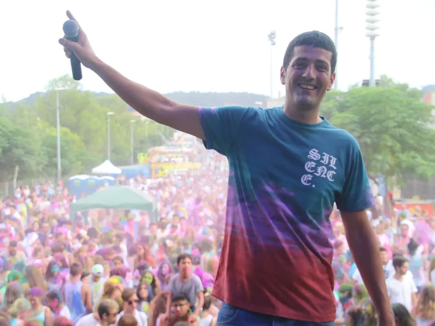 Animador de Fiesta Holi con peluca afro y micro señalando al publico cubierto de polvos de colores en plaza al aire libre