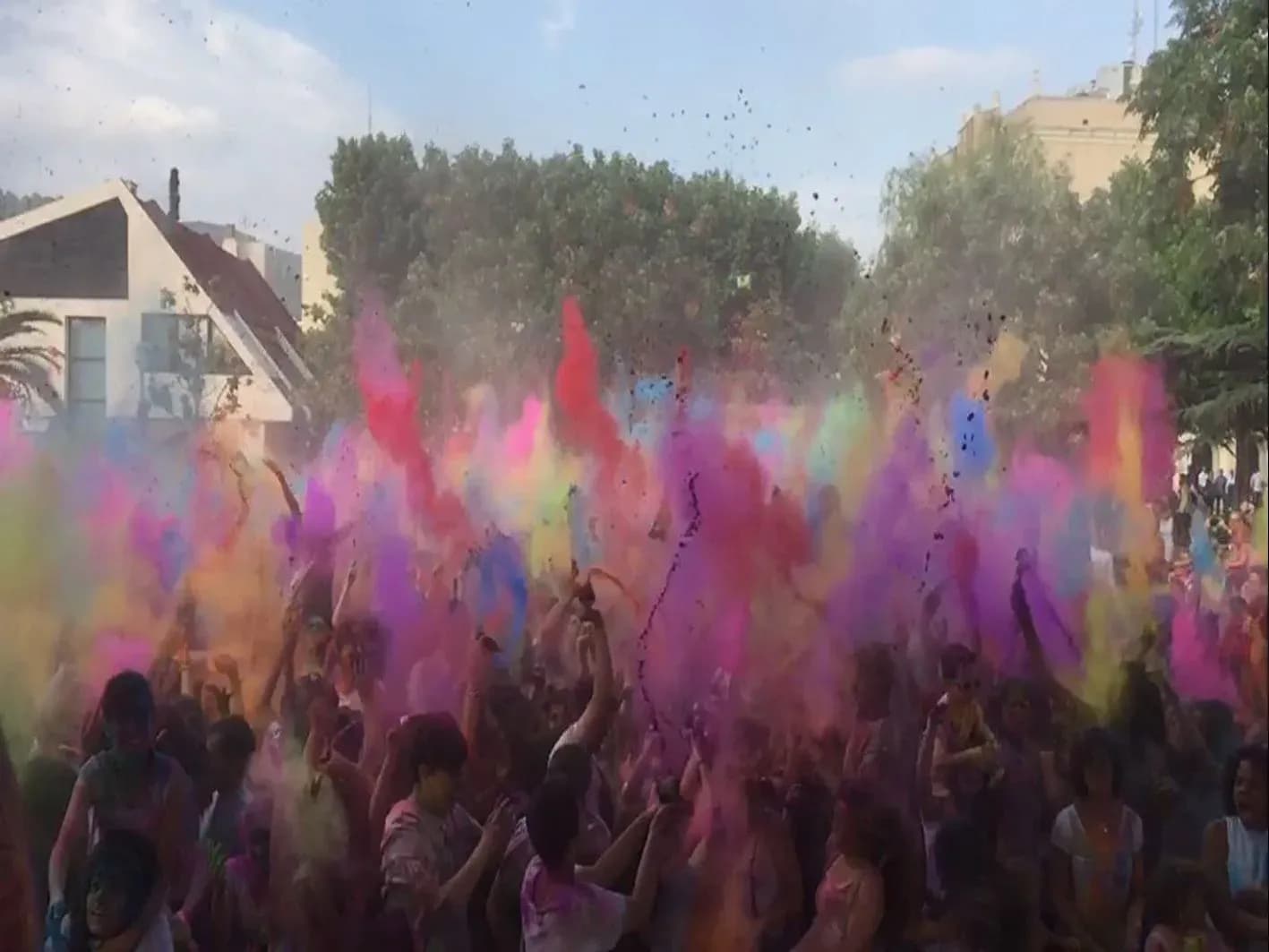 Multitud en fiesta Holi lanzando polvos de colores rojos, rosas y morados al aire entre arboles y casas
