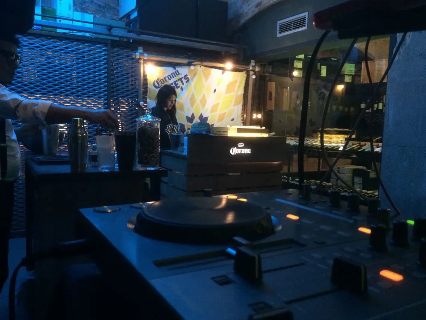 Cabina de DJ junto a barra Corona con coctelera y vinilo en plato, montaje para fiesta corporativa en local nocturno