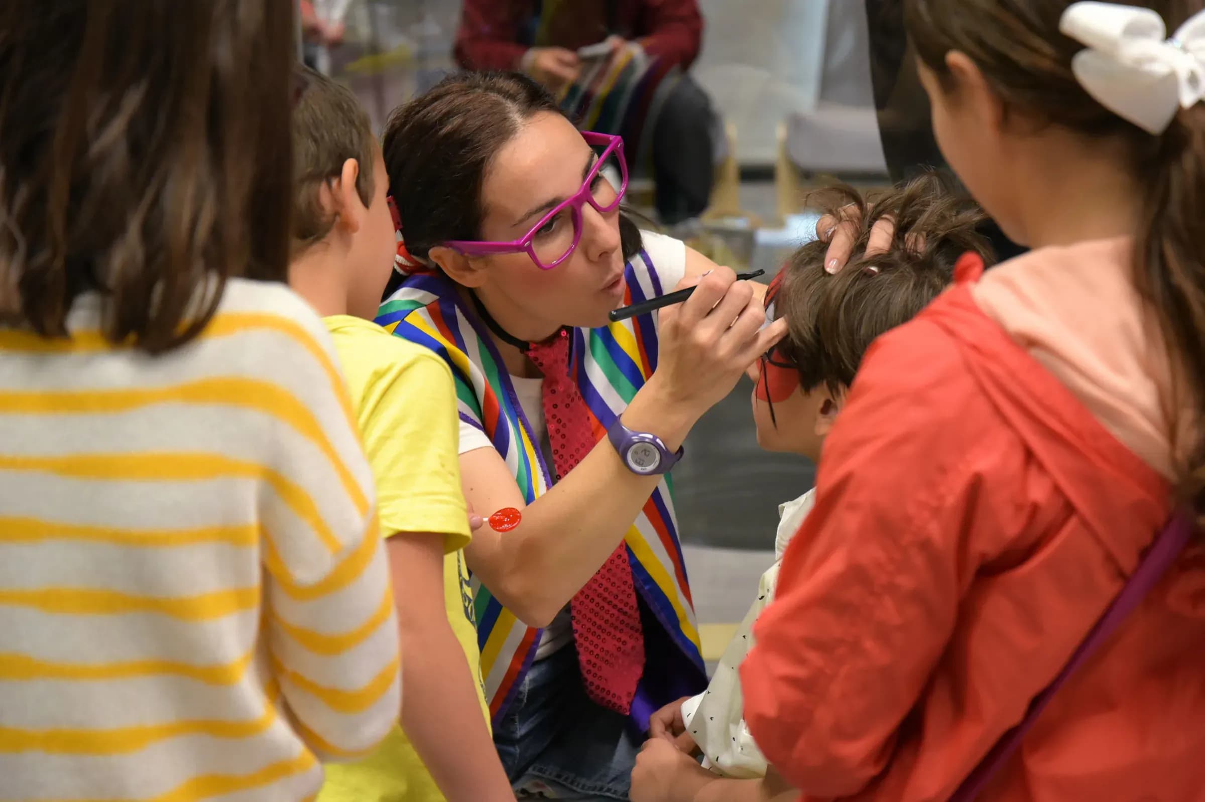 Animadora con gafas rosas pintando la cara de un nino en fiesta de empresa rodeada de otros ninos