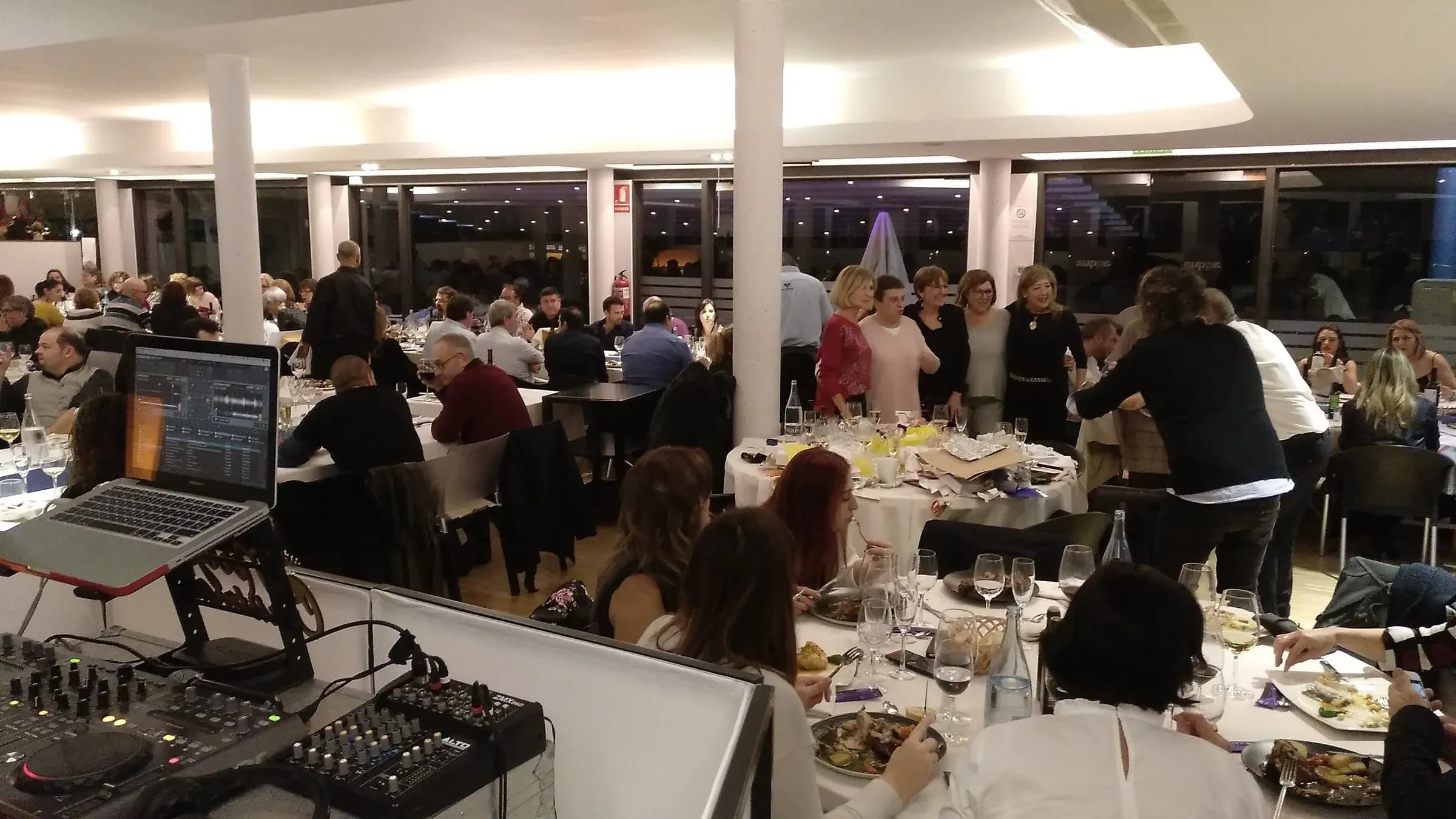 Cabina DJ con portatil y mesa de mezclas durante cena de fiesta de empresa con invitados en mesas y vistas nocturnas