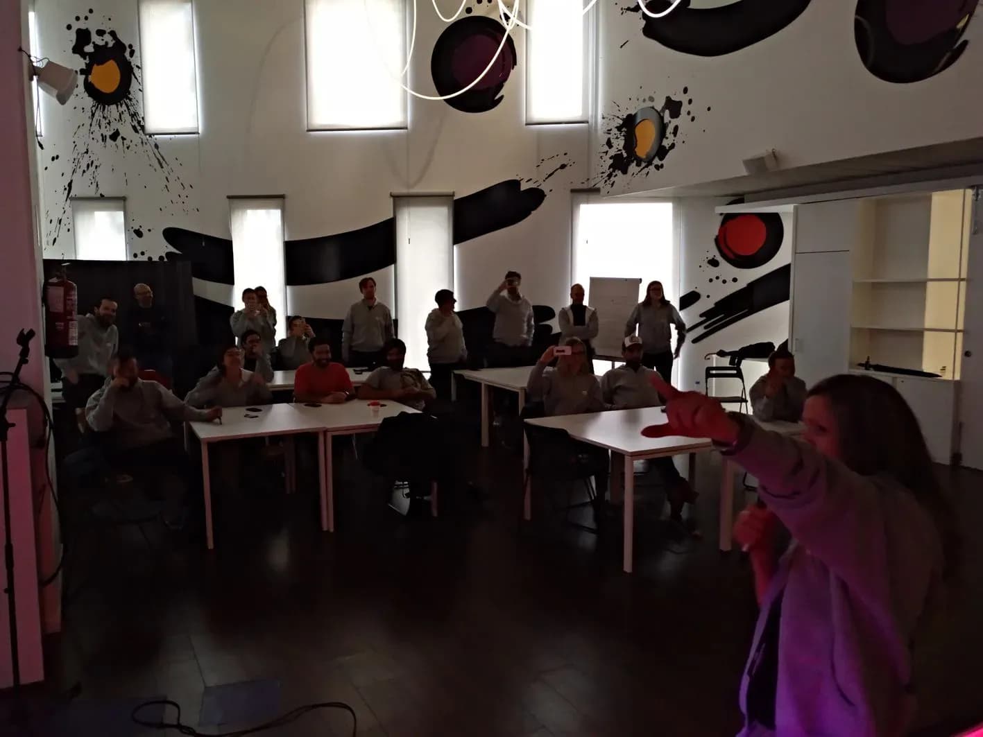 Fiesta de empresa en sala con paredes pintadas estilo manga, asistentes sentados en mesas y animadora gesticulando
