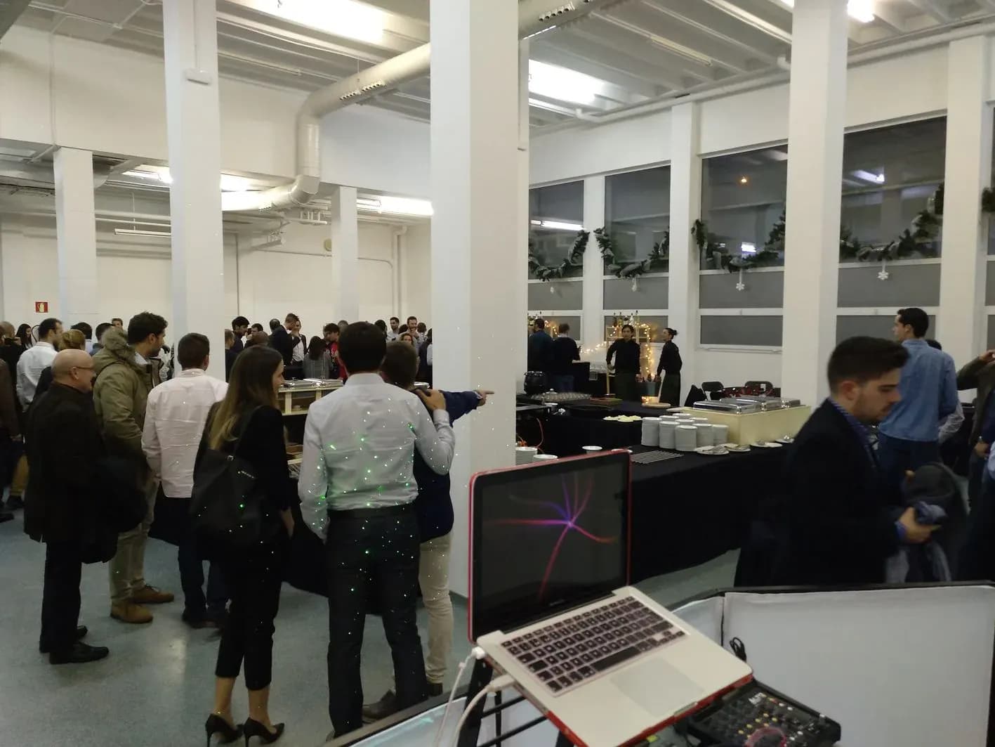 Cóctel de empresa con invitados conversando junto a buffet y MacBook con mesa de mezclas en sala industrial blanca