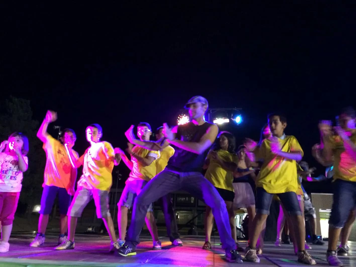 Animador con gorra bailando junto a niños con camisetas amarillas y naranjas sobre escenario con focos rosa y láseres