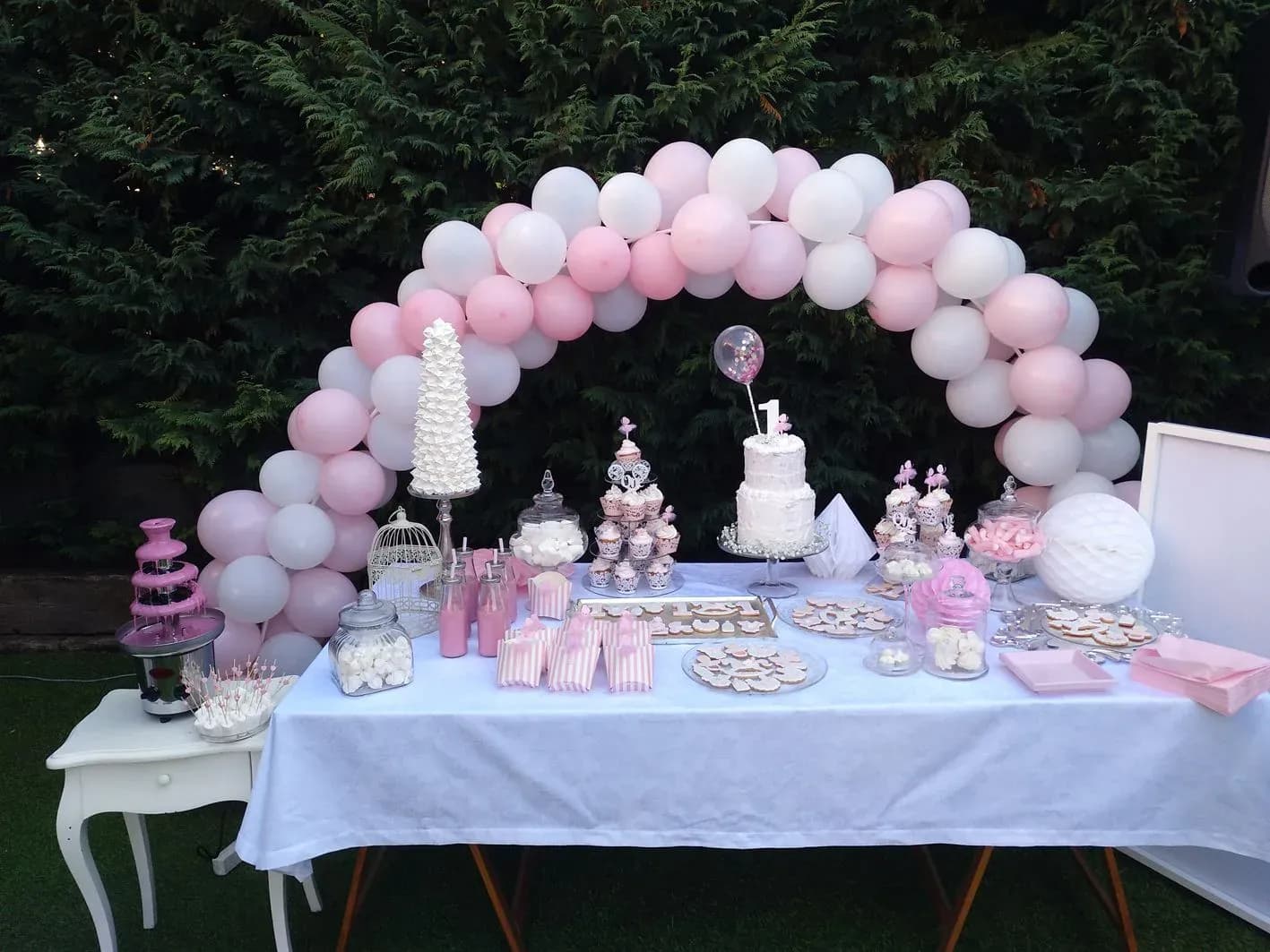 Mesa de chuches y pastel para fiesta infantil con arco de globos rosa y blanco, fuente de chocolate y candy bar al aire libre
