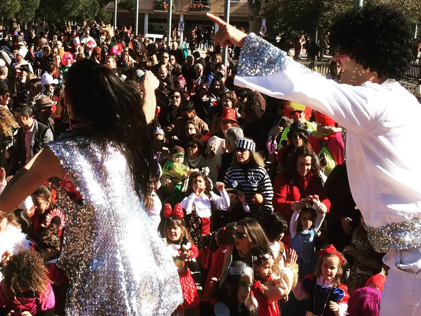 Animador con peluca rizada blanca sobre escenario señalando a niños disfrazados de carnaval entre publico familiar