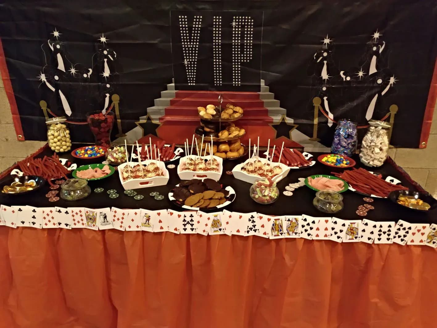Mesa de candy bar tematica VIP con bandejas de dulces, frutos secos y naipes decorativos sobre mantel rojo y negro