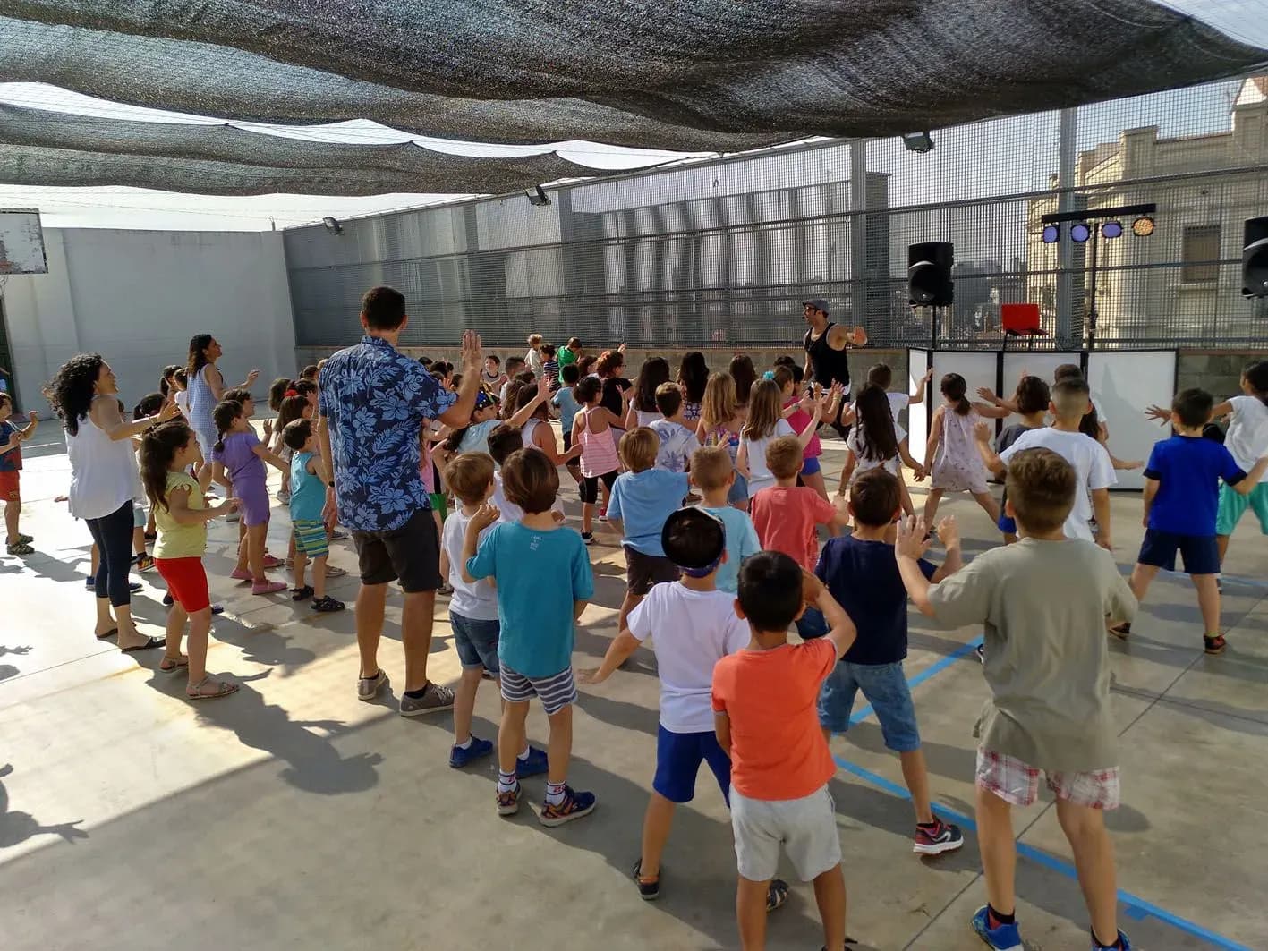Animadores dirigiendo coreografia infantil en pista exterior con grupo numeroso de ninos bailando bajo toldo