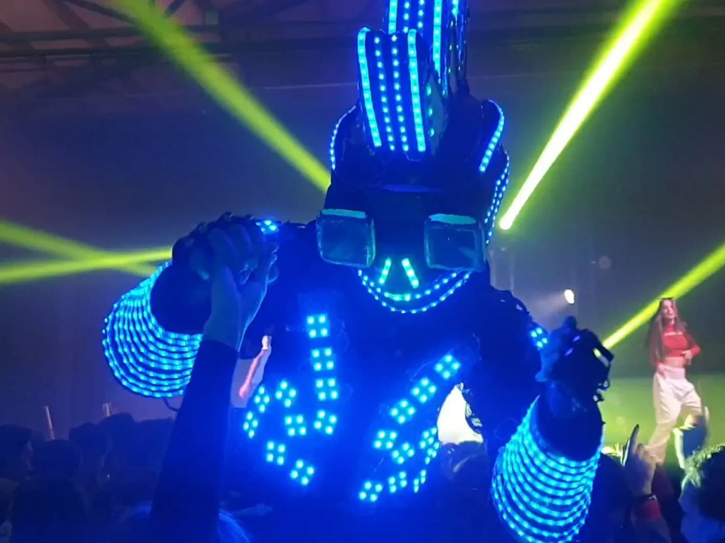 Robot LED azul con cresta luminosa y gafas tubulares de pie entre la multitud durante el show de un fiestón con láseres