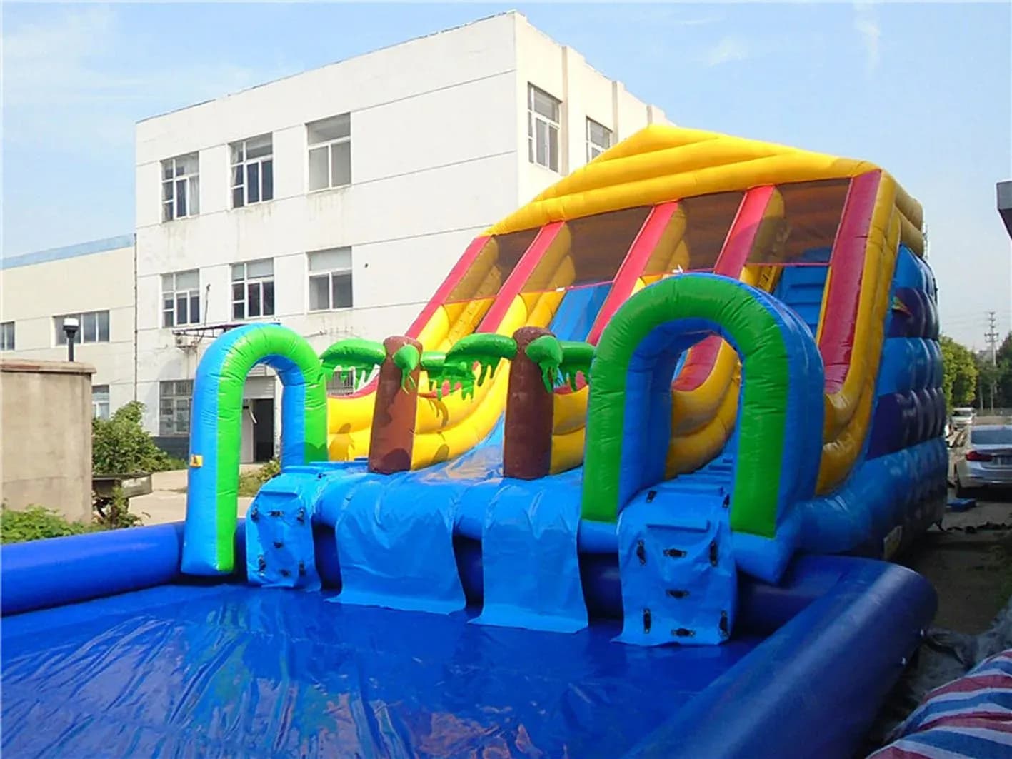 Tobogán hinchable acuático azul, amarillo y rosa con piscina y rampas frente a fábrica, modelo XL para fiestas estivales