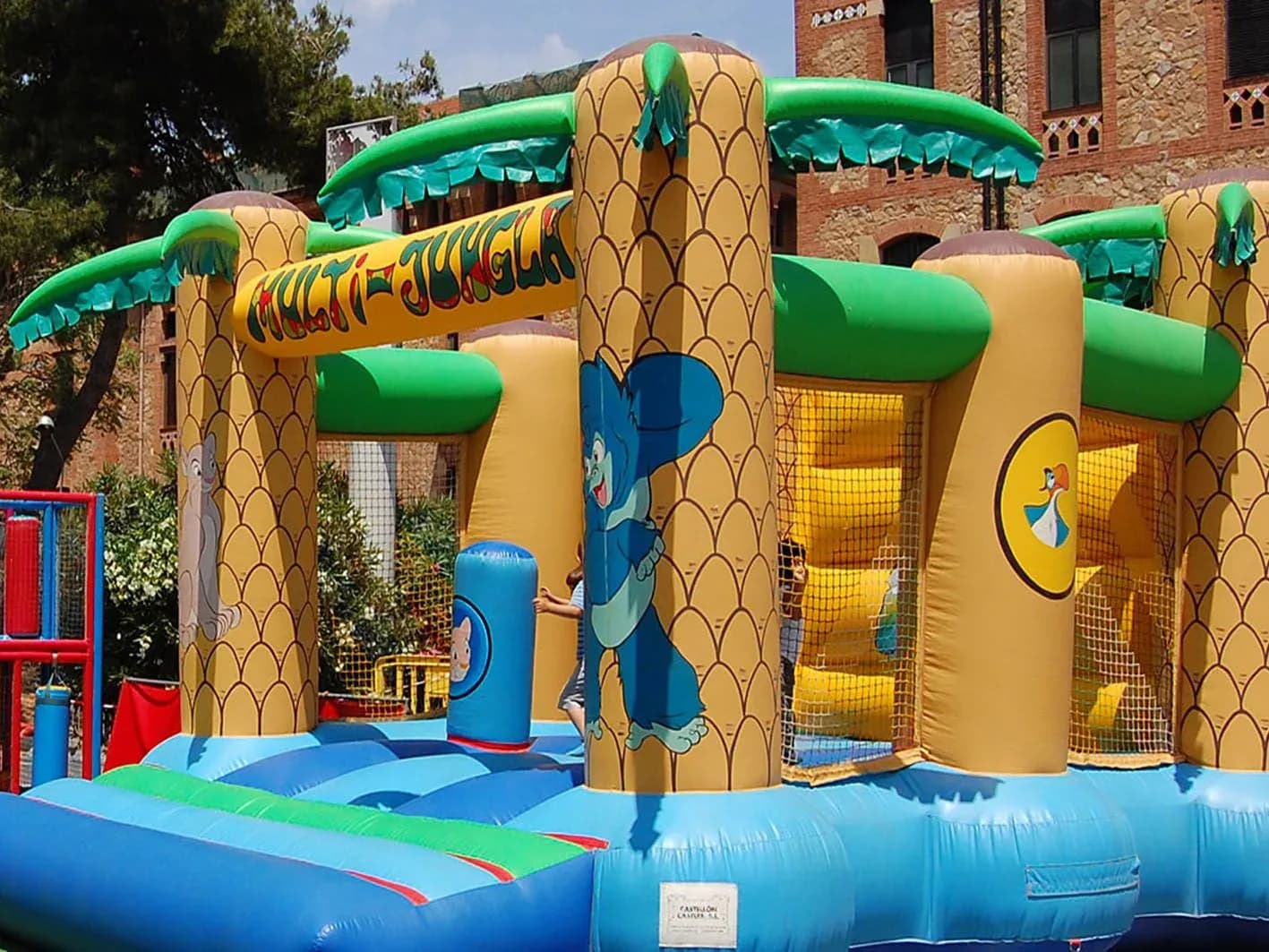 Multi-aventura hinchable Multi-Jungla con palmeras, animales y red de escalada en plaza junto a edificio de ladrillo