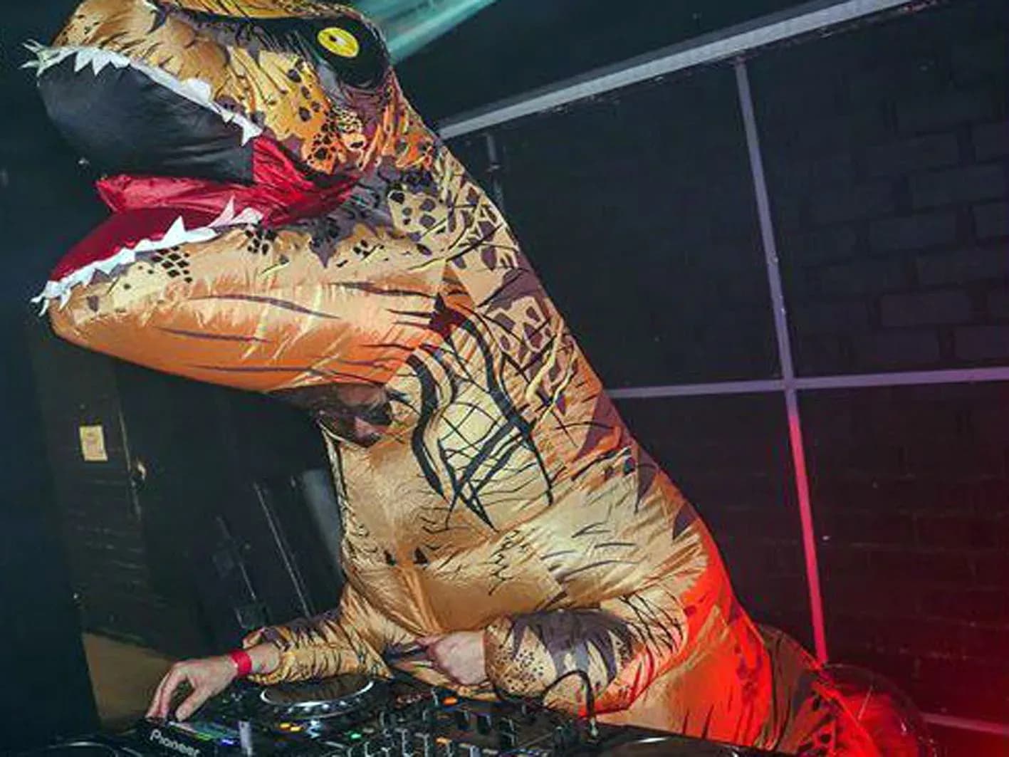 DJ disfrazado de T-Rex hinchable marrón inclinado sobre la mesa Pioneer ante muro de ladrillo, sesión de Hunder & Button