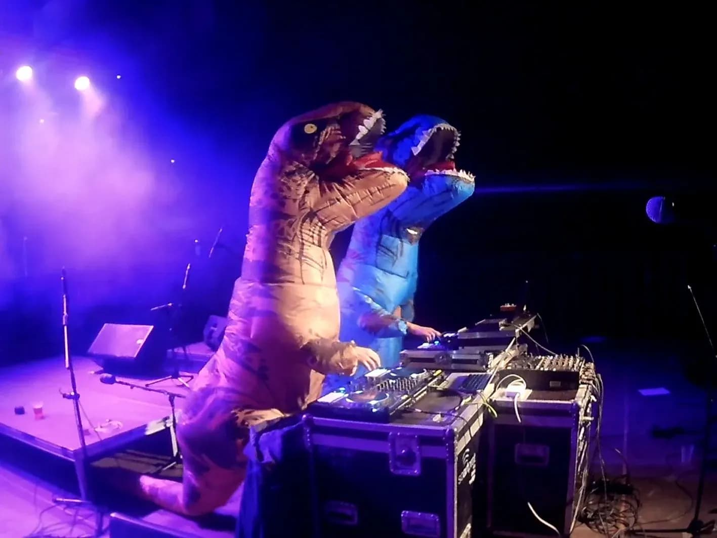 Dúo Hunder & Button con trajes inflables de tiranosaurio sobre escenario con focos morados y mesa de DJ profesional