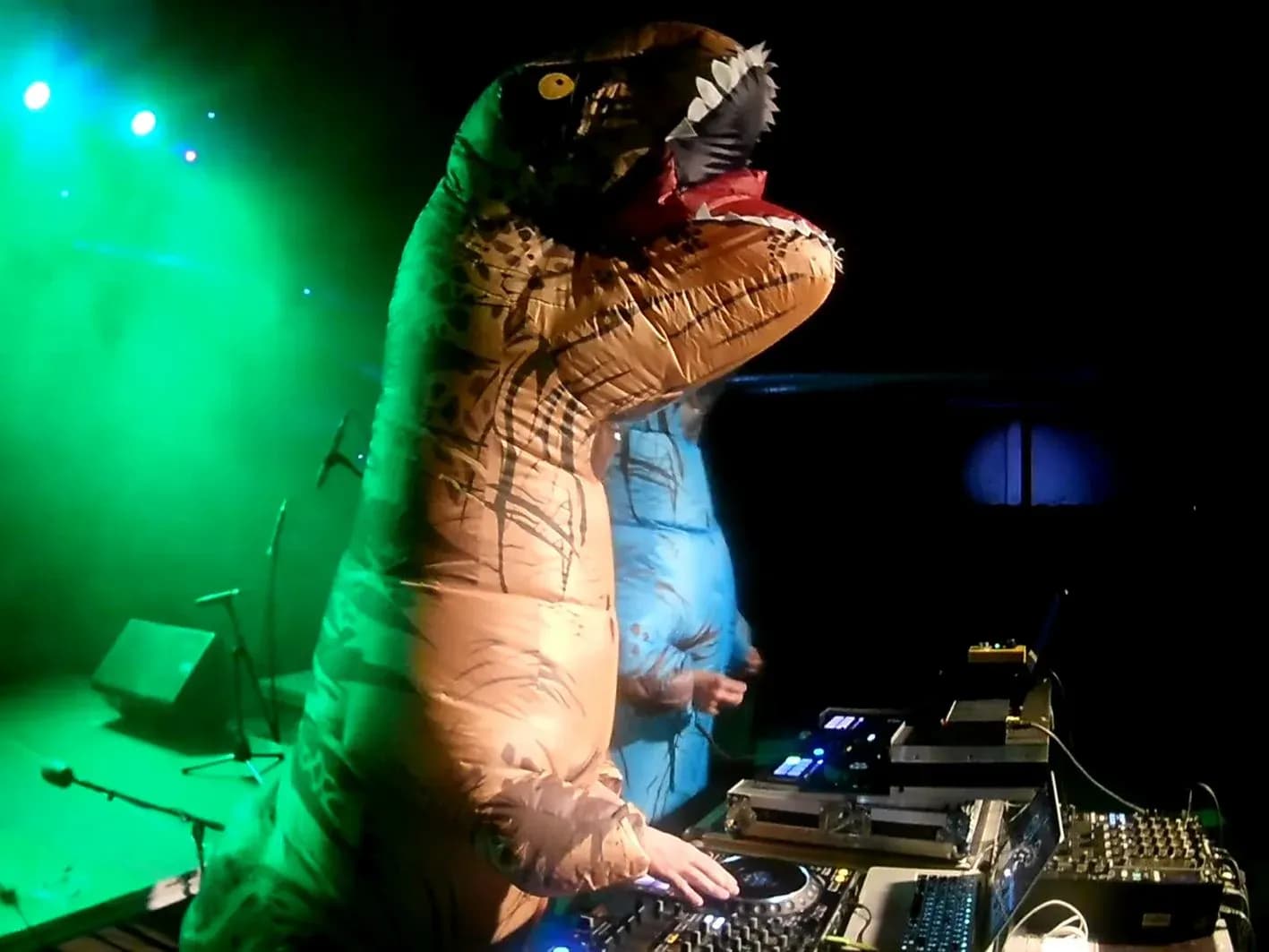 DJ disfrazado de T-Rex hinchable manipulando los CDJ Pioneer bajo luz verde escénica, espectáculo de Hunder & Button