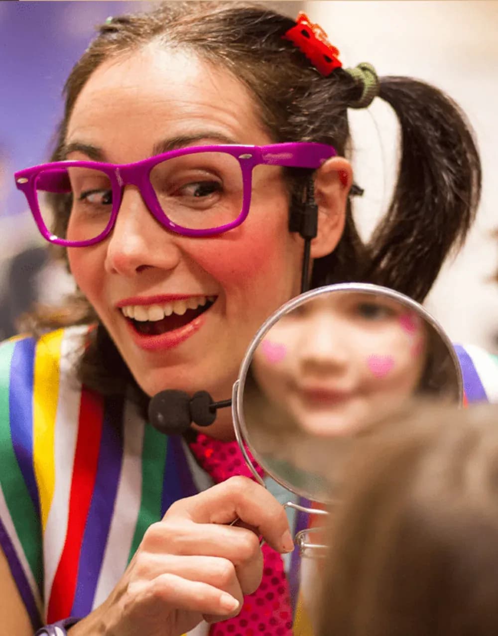 Jessica con gafas magenta y micro vincha sosteniendo espejo redondo donde se ve una niña con maquillaje de fantasía rosa