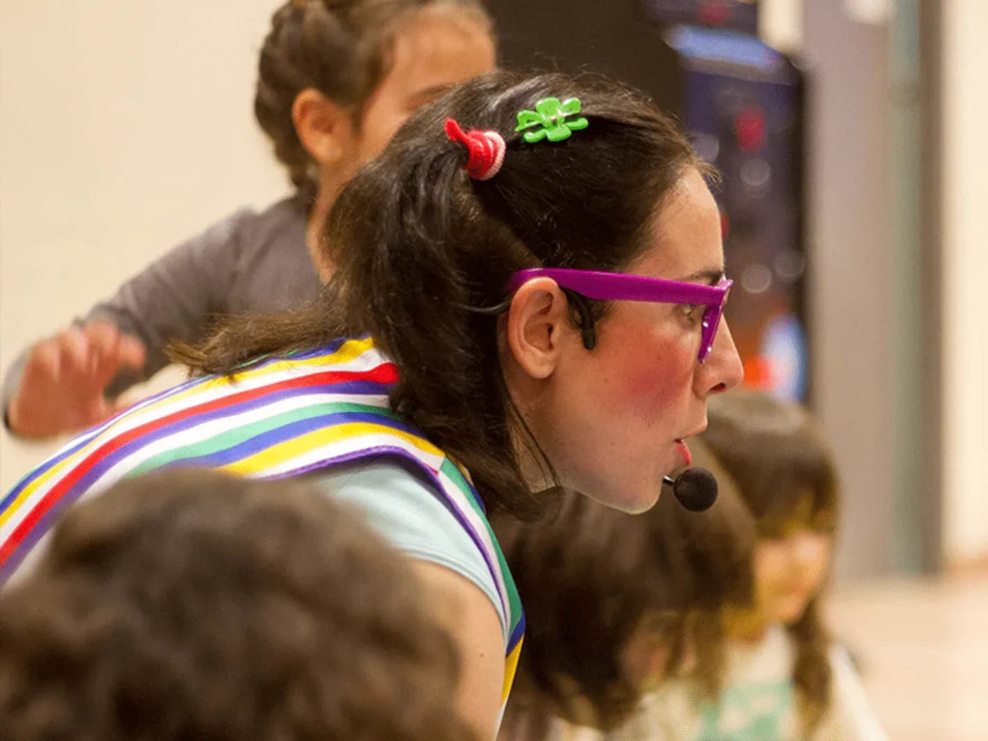 Jessica con micro vincha y chaleco arcoíris inclinada sobre el grupo de niños durante animación infantil en sala de eventos