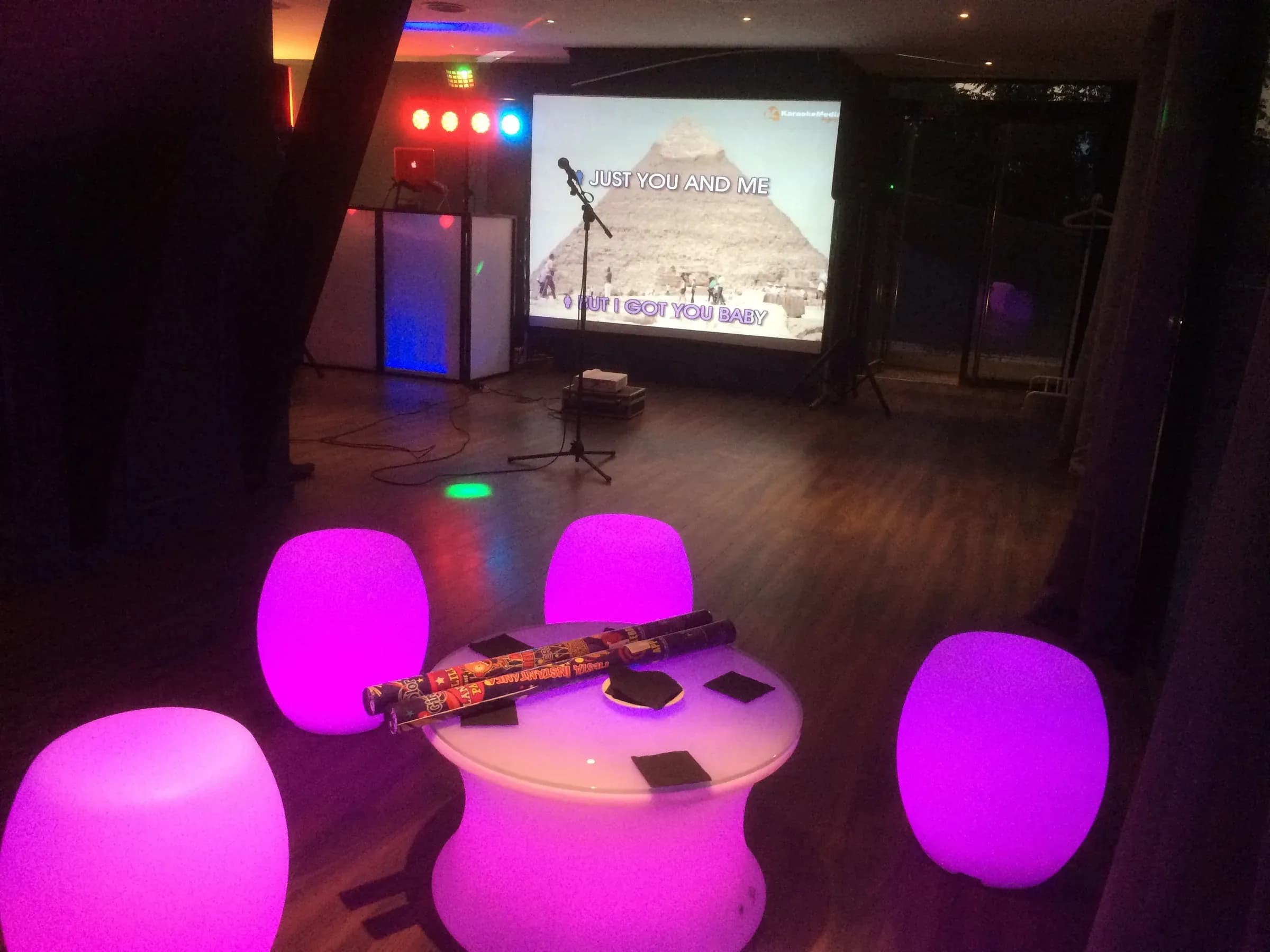 Set de karaoke con pantalla, micro en pie y taburetes LED rosa con mesa baja y luces de discoteca en restaurante