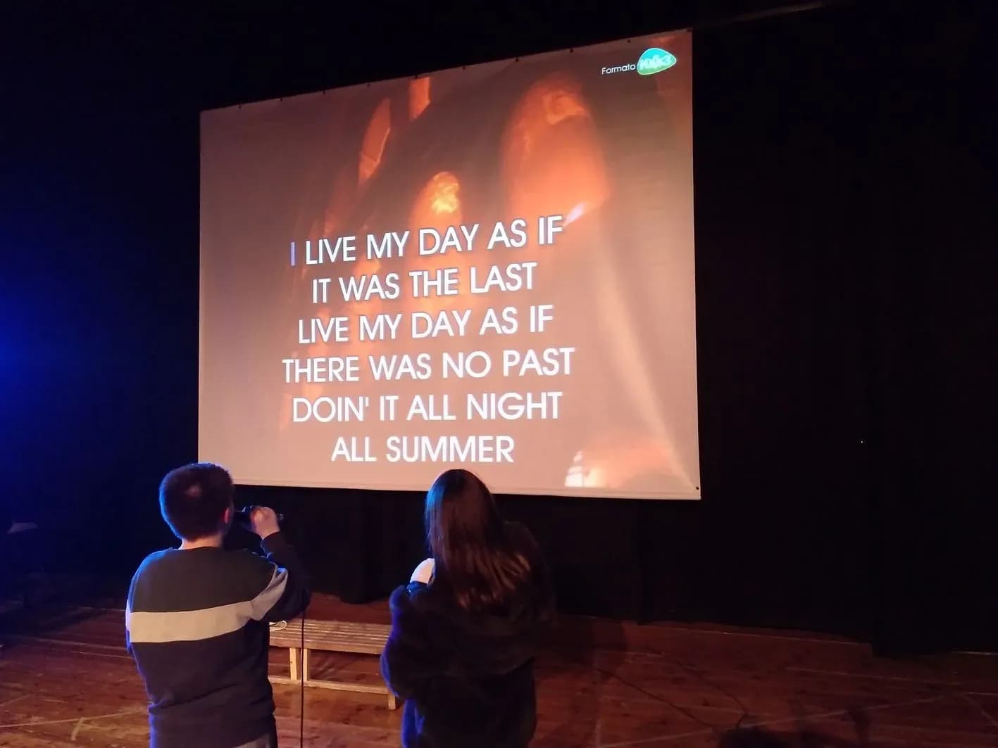 Pareja de jovenes de espaldas cantando karaoke frente a pantalla con letra de cancion en ingles en sala oscura