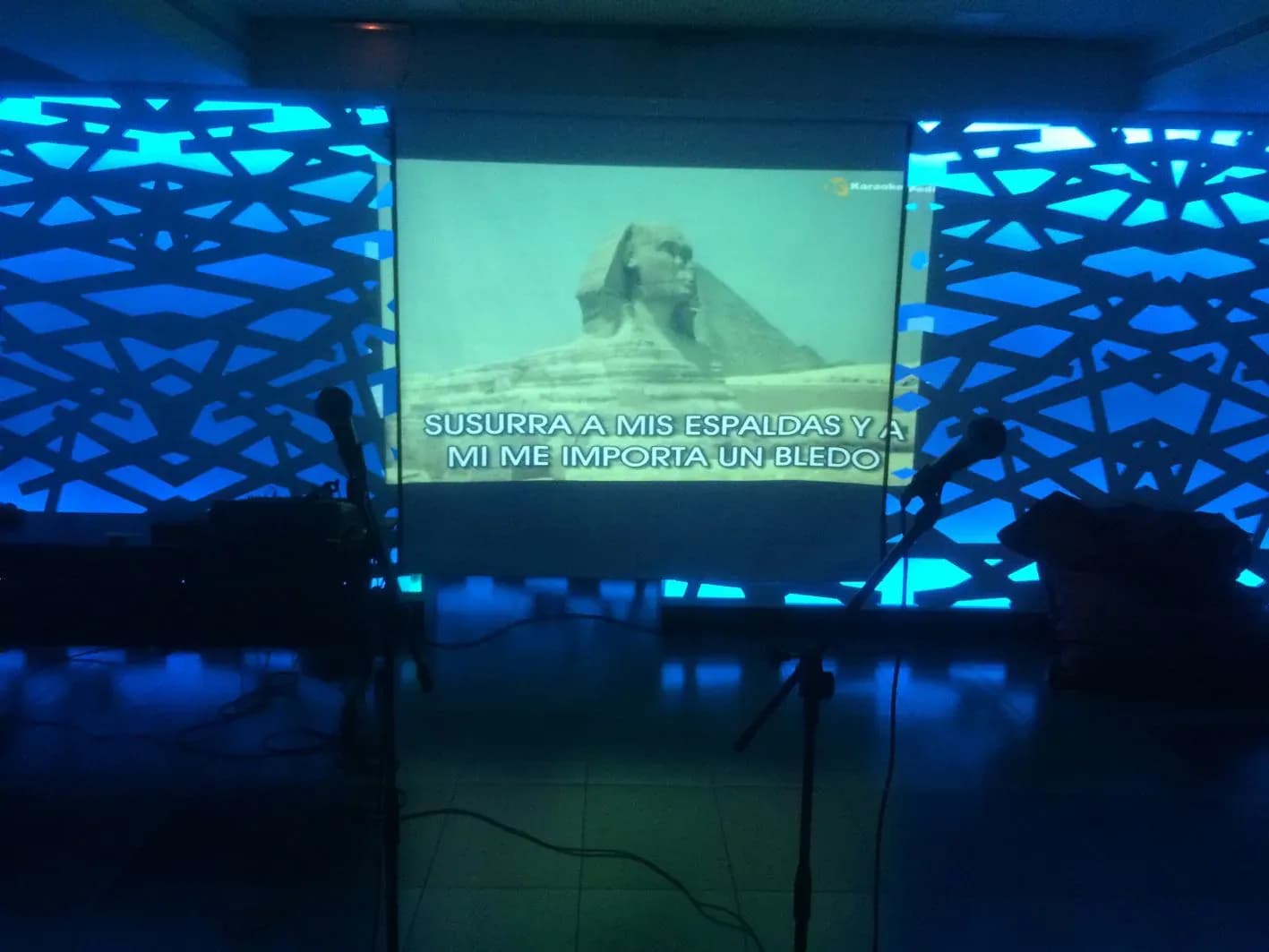 Sala de karaoke con pantalla proyectando letra de cancion y esfinge, paneles azules iluminados y dos microfonos