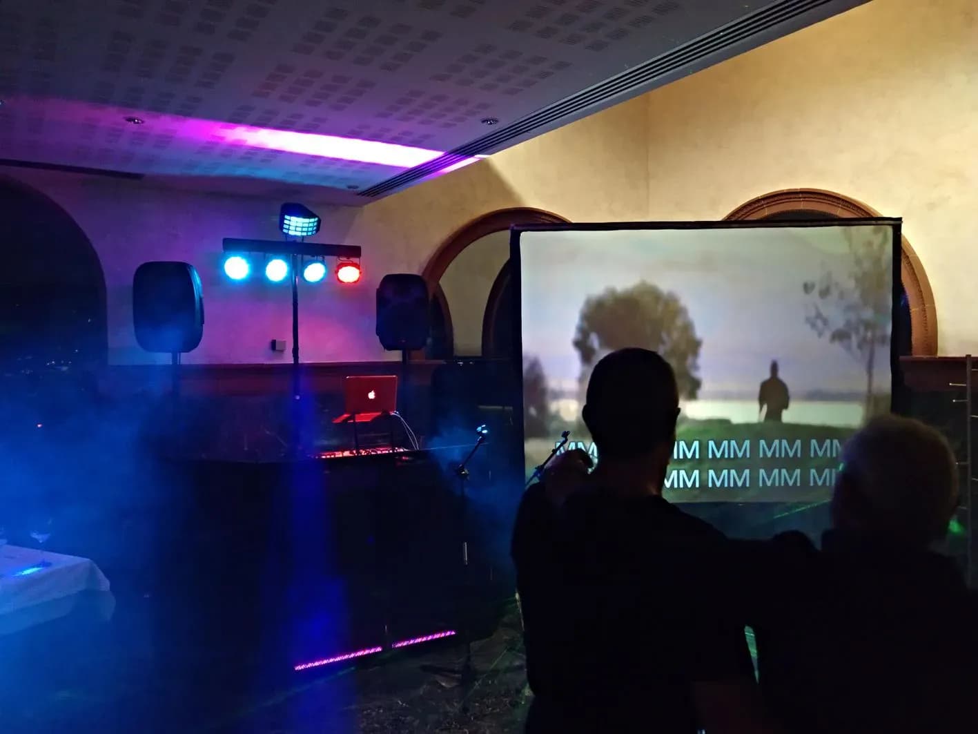 Sesion de karaoke en salon de eventos con pantalla, focos de colores y publico cantando entre humo